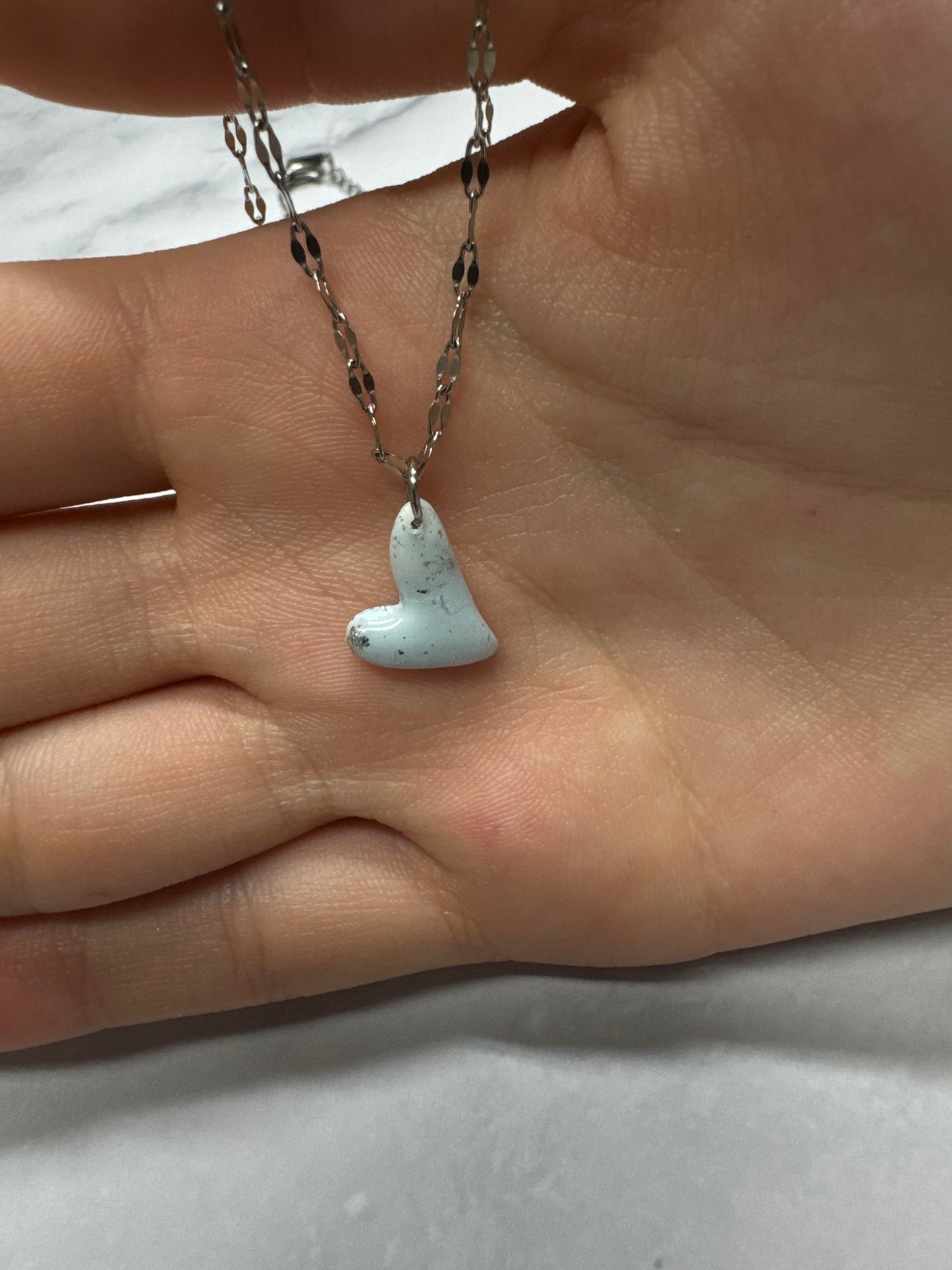 Ciondolo a cuore azzurro tenue appoggiato sul palmo di una mano, catena argentata.
