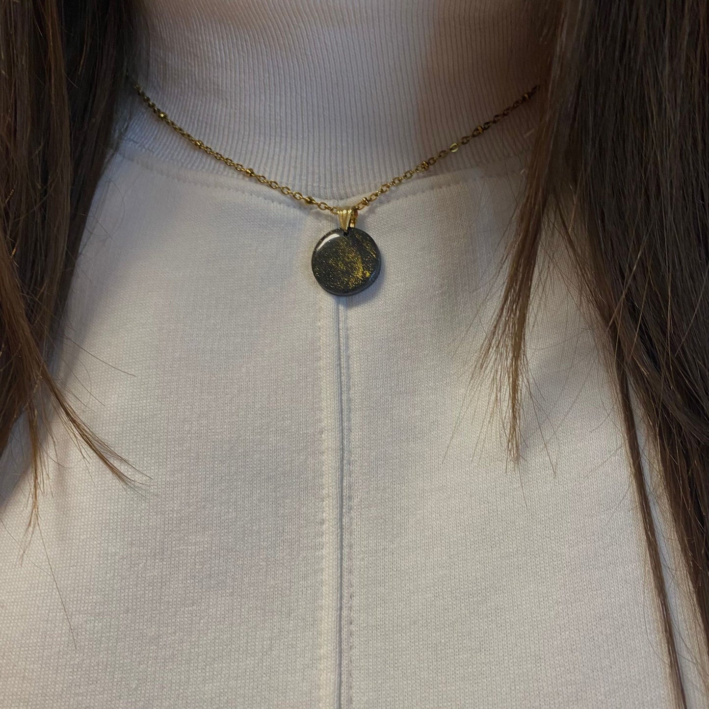 Primo piano della collana indossata su una maglietta bianca. Collana pendente con ciondolo rotondo in porcellana fredda, colore nero con dettagli in oro, con catenina dorata.
Gioiello artigianale fatto a mano da persone cieche e ipovedenti.