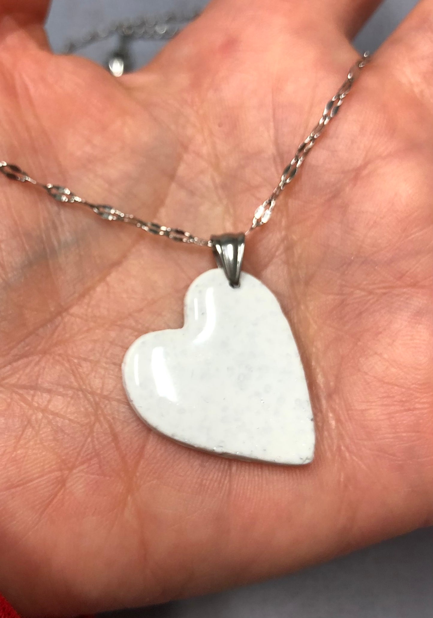 Mano che tiene nel palmo una collana con ciondolo a cuore bianco a pois argentati in porcellana fredda bianca, sfumature dorate e catenina argentata. Gioiello artigianale fatto a mano da persone cieche e ipovedenti.