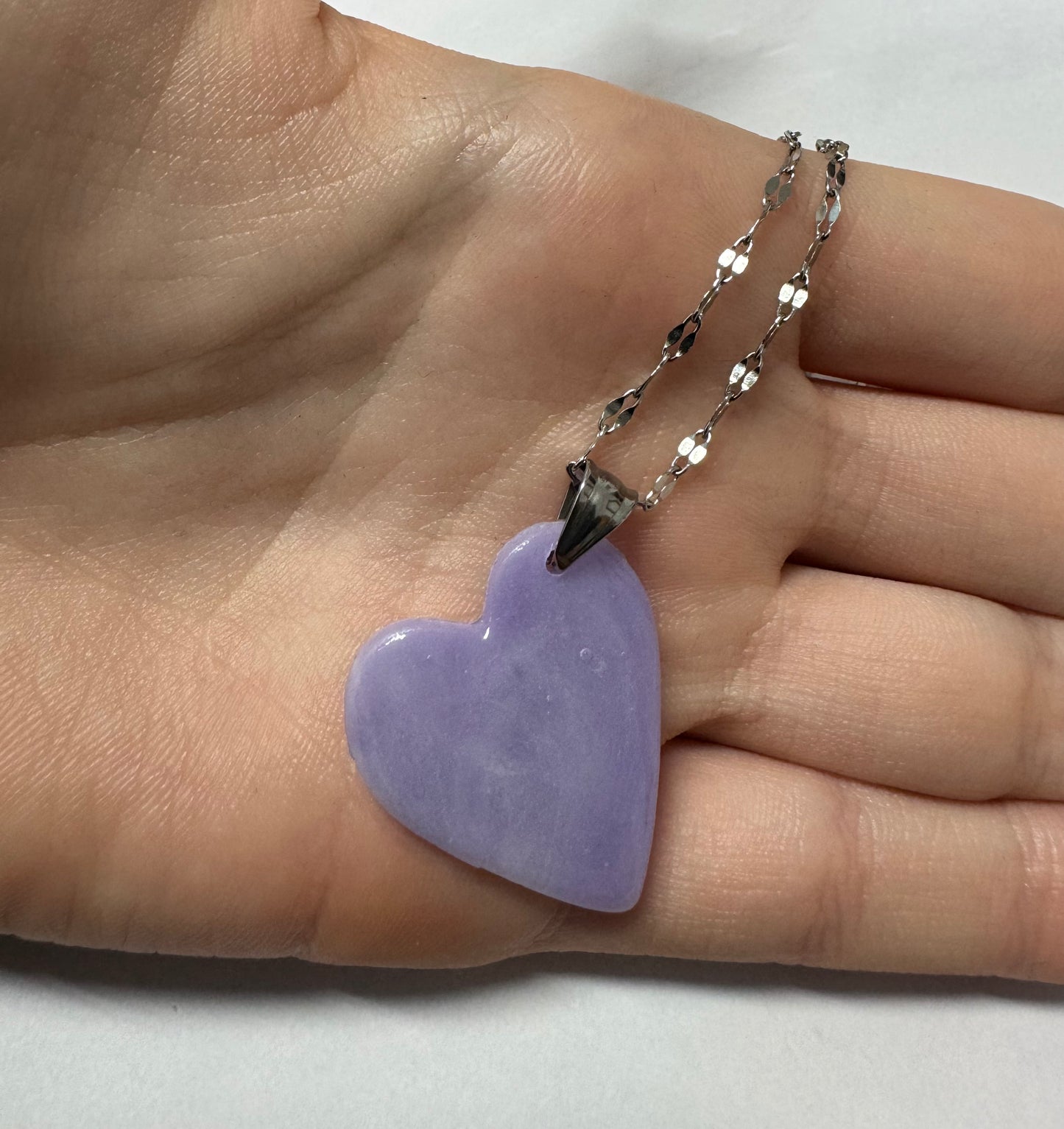 Mano che tiene nel palmo una collana con ciondolo a cuore lilla in porcellana fredda e catenina argentata. Gioiello artigianale fatto a mano da persone cieche e ipovedenti.