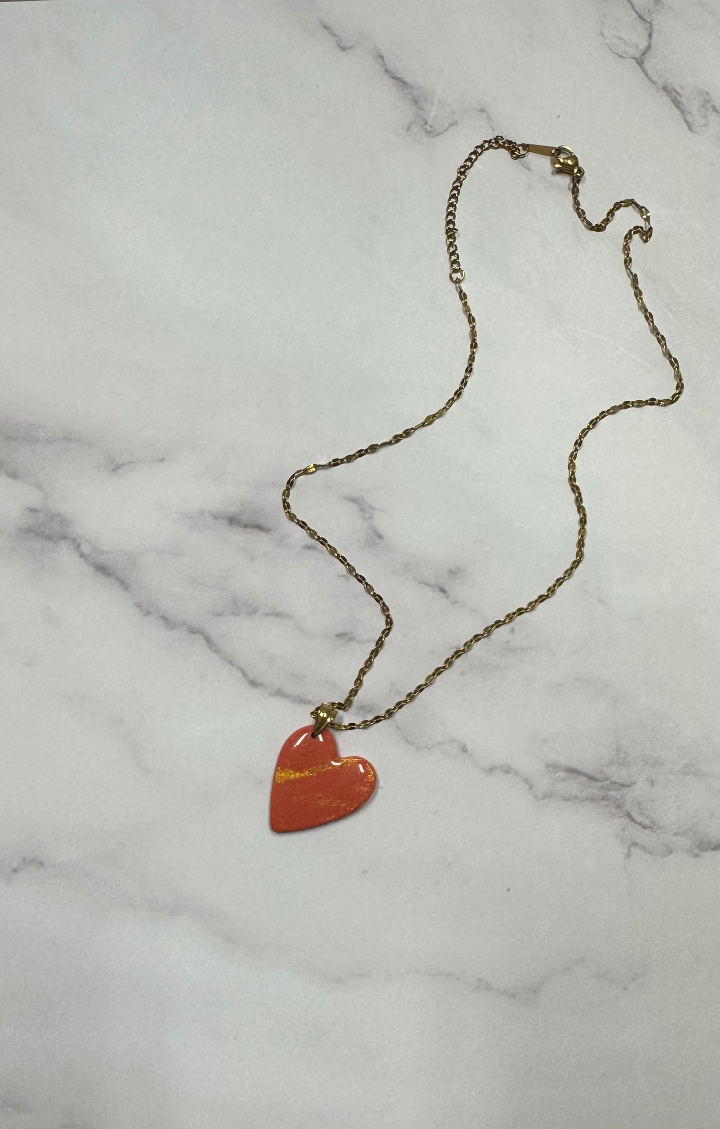 Collana dorata con ciondolo a cuore rosso in porcellana fredda e venature oro, adagiata su fondo marmoreo. Creata da artigiani ciechi e ipovedenti.