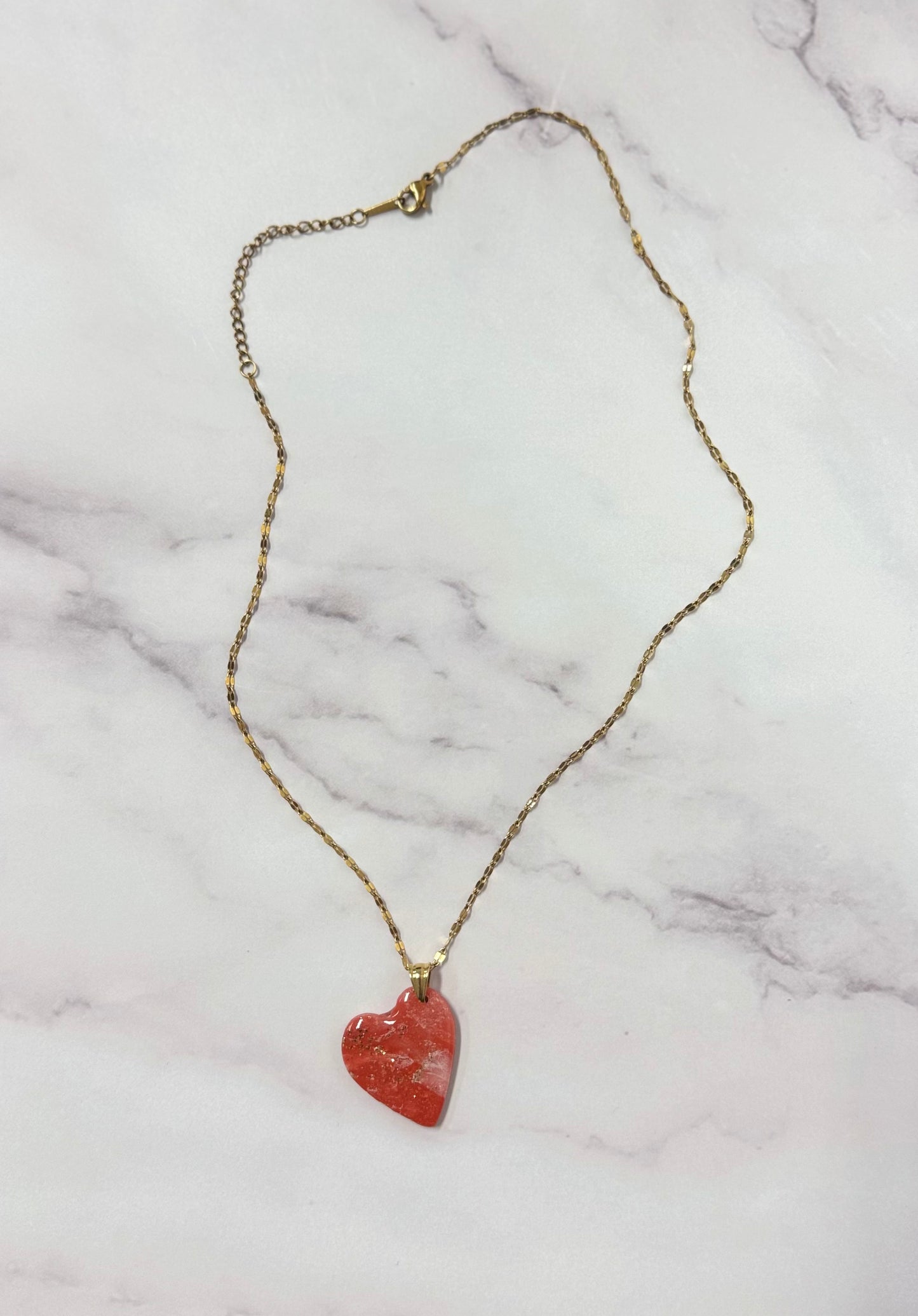Collana dorata con ciondolo a cuore rosso marmorizzato in porcellana fredda e sfumature dorate, adagiata su fondo marmoreo. Creata da artigiani ciechi e ipovedenti
