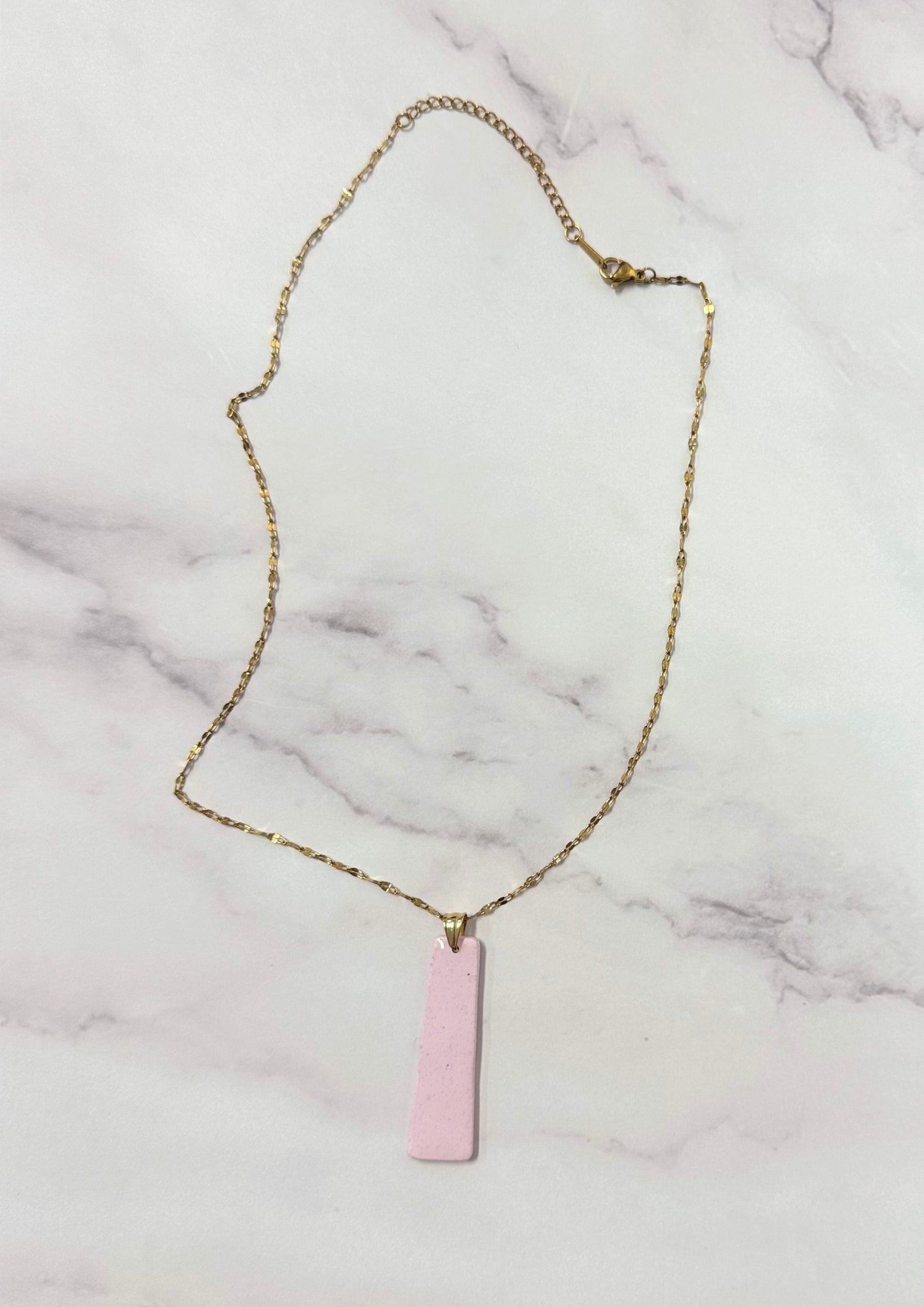 Collana dorata con ciondolo a forma di rettangolo, rosa, in porcellana fredda, adagiata su fondo marmoreo. Creata da artigiani ciechi e ipovedenti.