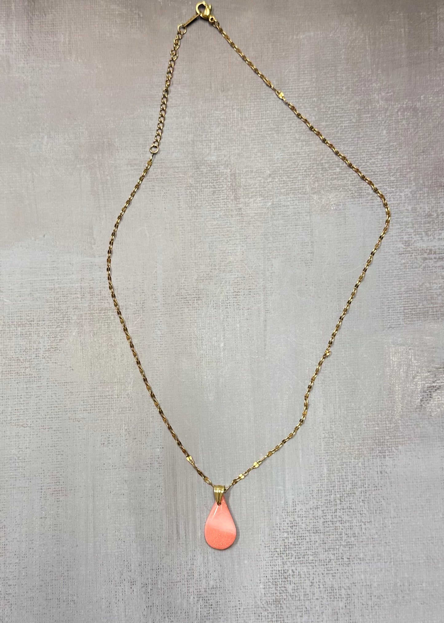 Collana dorata con ciondolo a goccia rosa corallo in porcellana fredda, adagiata su fondo grigio. Creata da artigiani ciechi e ipovedenti.