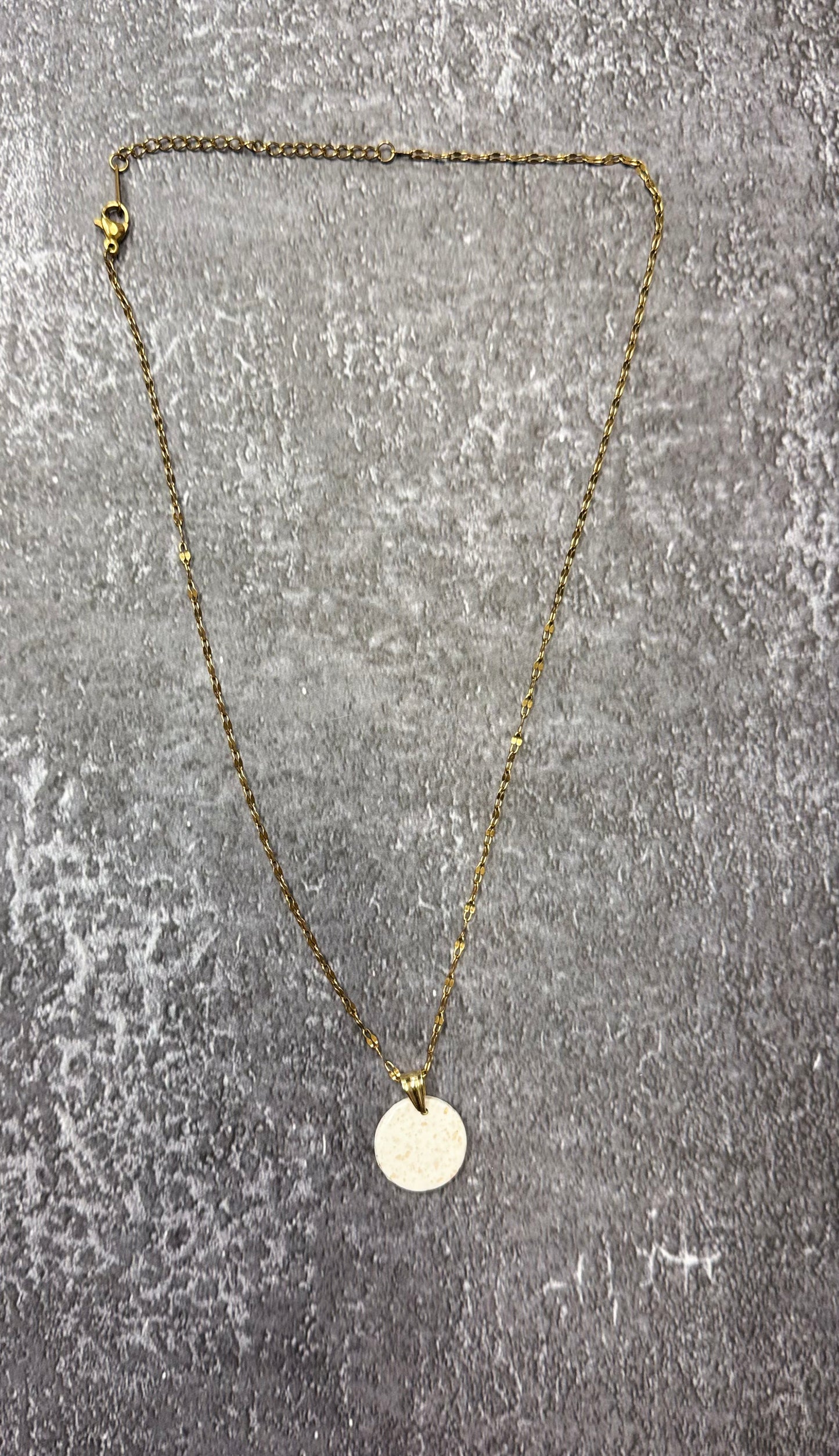 Collana con catenina dorata e pendente tondo bianco con pallini dorati in porcellana fredda e glitter, adagiata su fondo marmoreo. Creata da artigiani ciechi e ipovedenti.