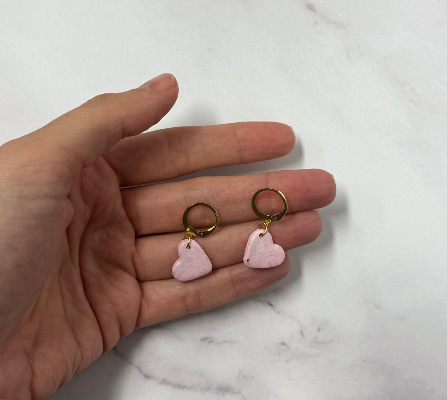 Orecchini con cerchi dorati e ciondolo a forma di cuore rosa in porcellana fredda, sul palmo di una mano. Gioiello realizzato da artigiani ciechi e ipovedenti