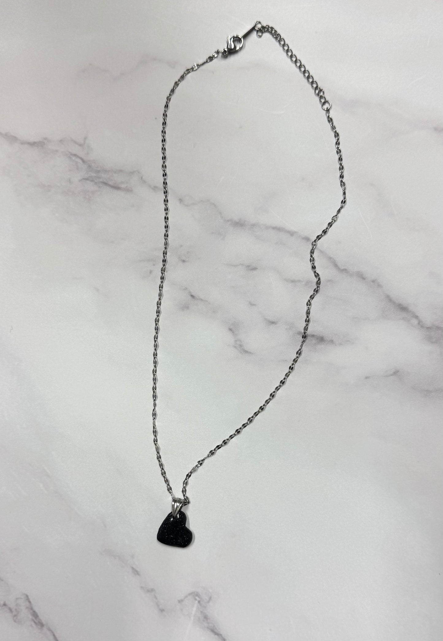 Collana cuoricino nero con brillantini argento, catena intera adagiata su sfondo marmo.
