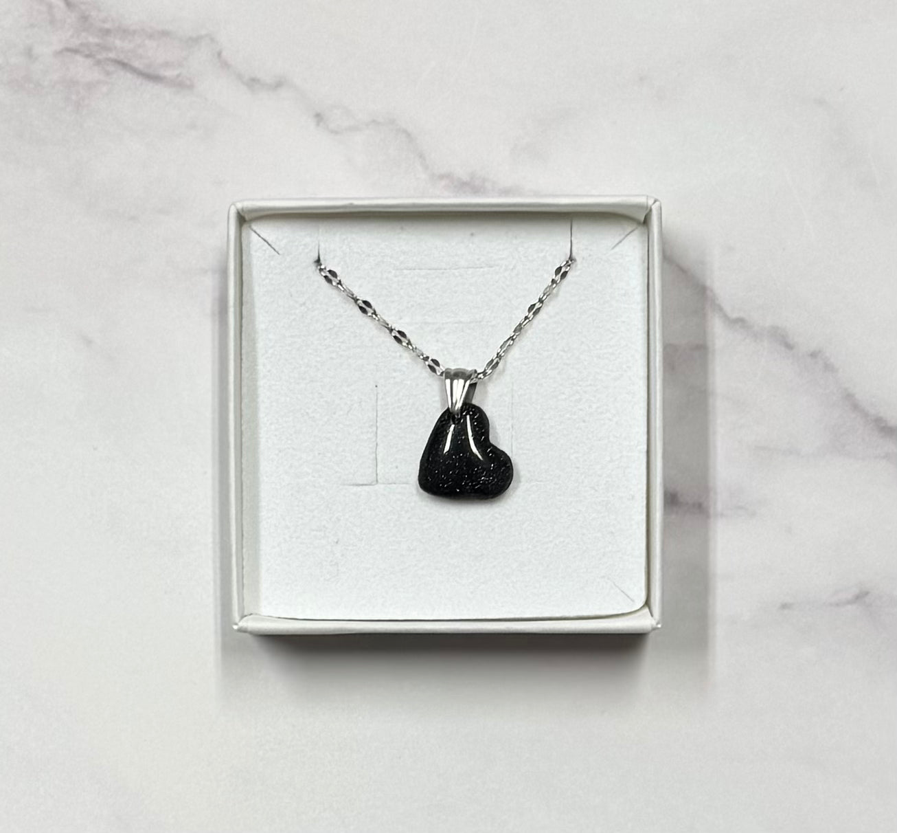 Collana cuoricino nero con brillantini argento, in scatolina regalo bianca aperta su sfondo marmo.