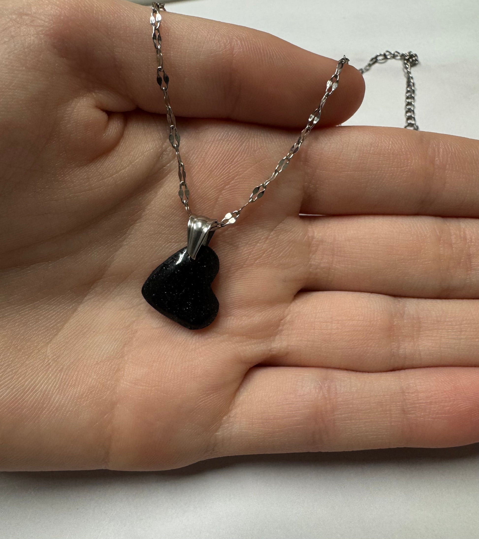 Ciondolo a cuore nero con brillantini argento su catena d'argento, appoggiato sul palmo di una mano.