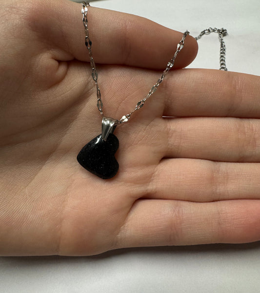 Ciondolo a cuore nero con brillantini argento su catena d'argento, appoggiato sul palmo di una mano.