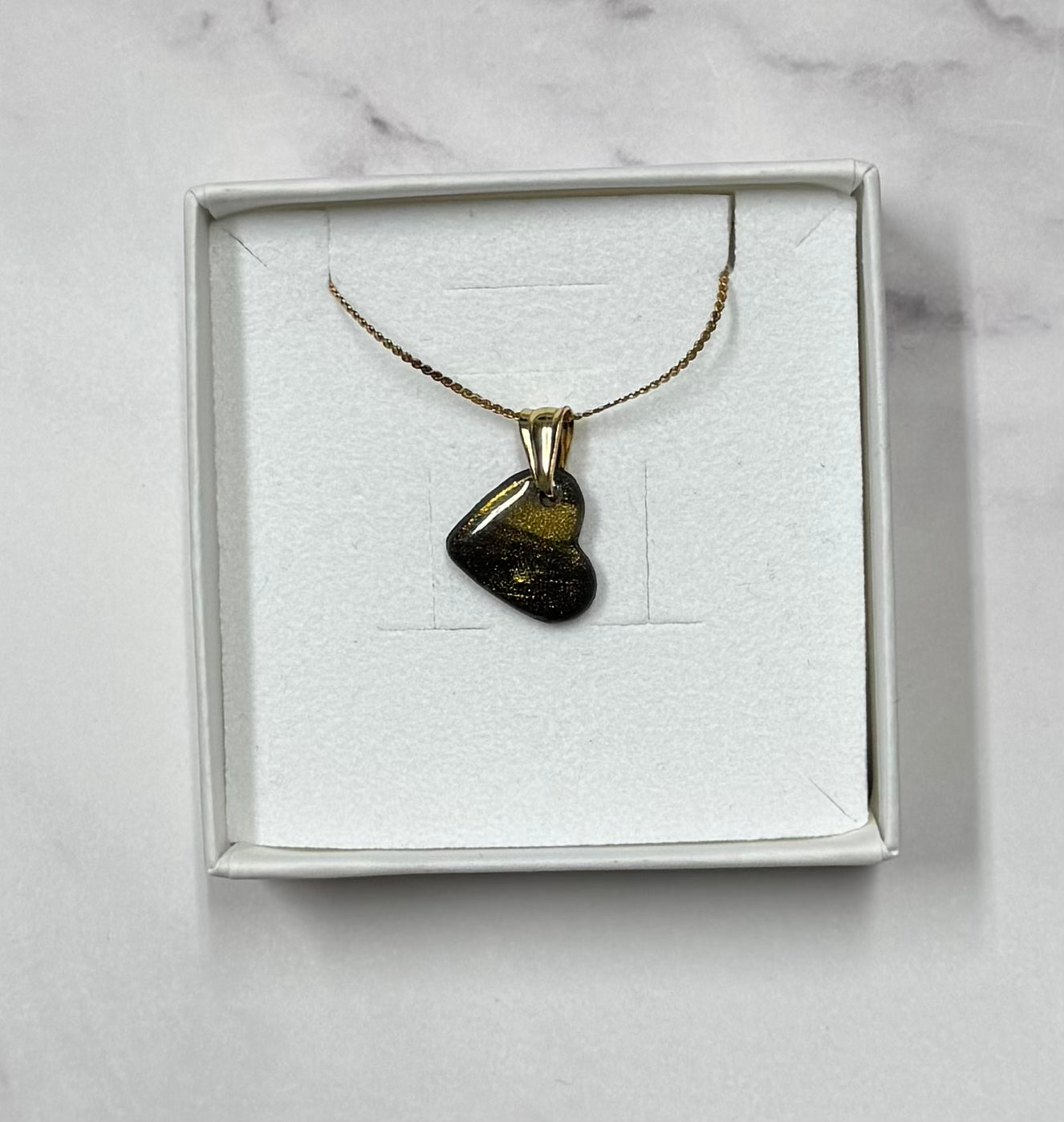 Collana cuoricino nero con brillantini oro, in scatolina regalo bianca aperta su sfondo marmo.