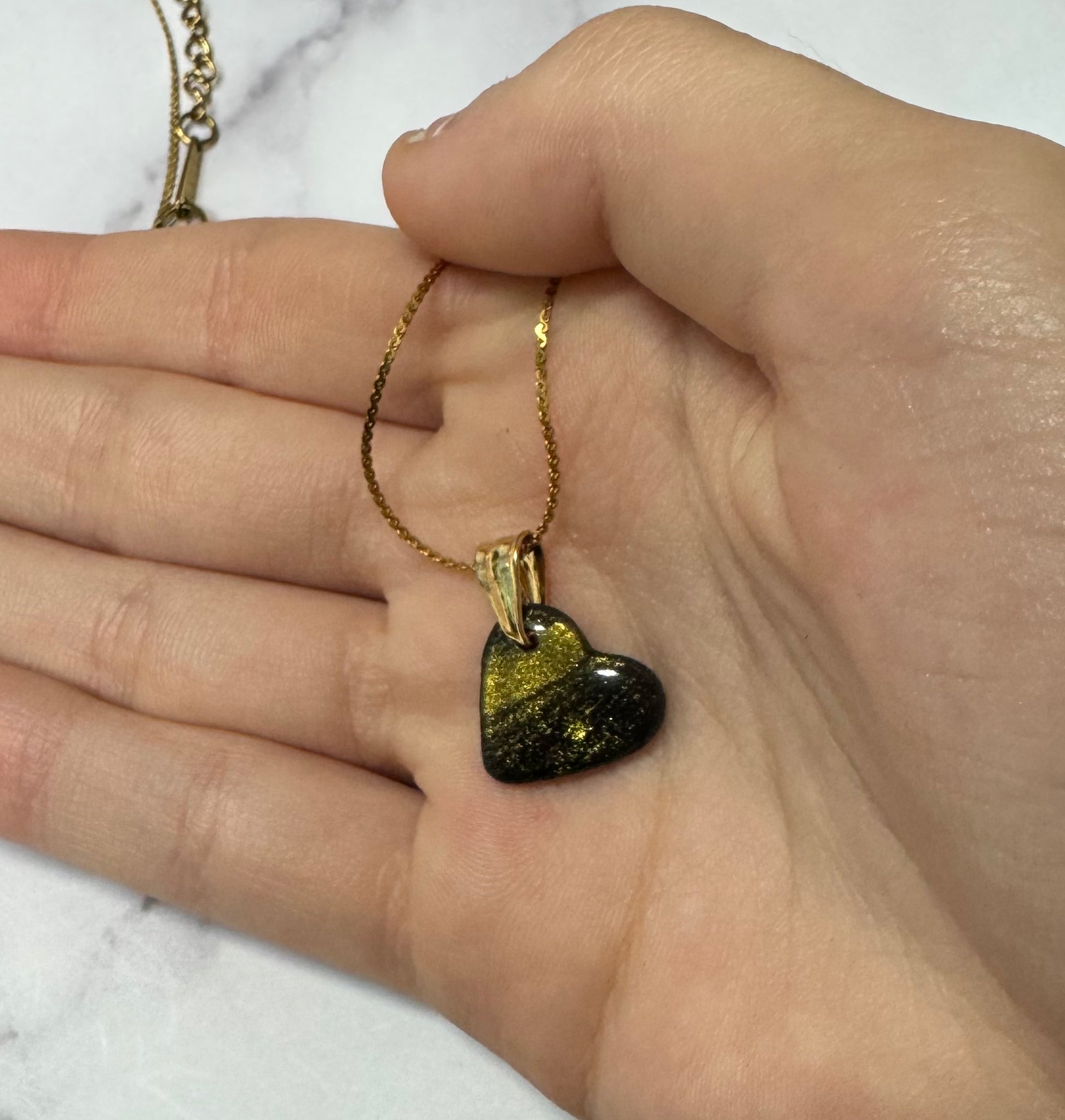 Ciondolo a cuore nero con brillantini oro, tenuto tra le dita di una mano, catena dorata.