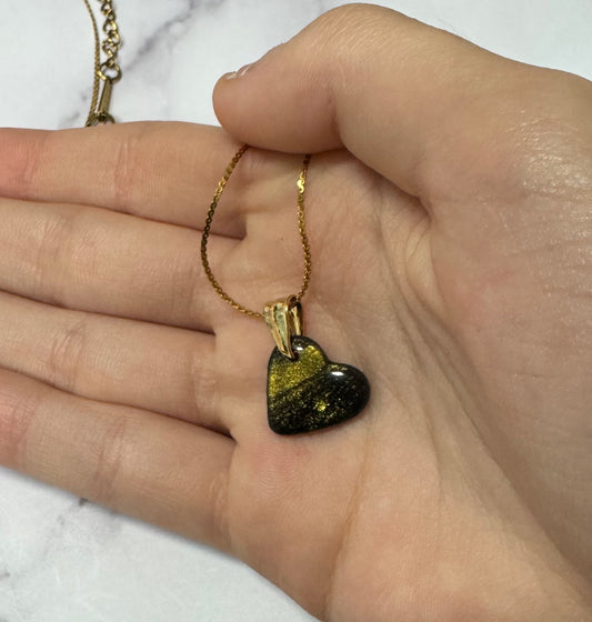 Ciondolo a cuore nero con brillantini oro, tenuto tra le dita di una mano, catena dorata.