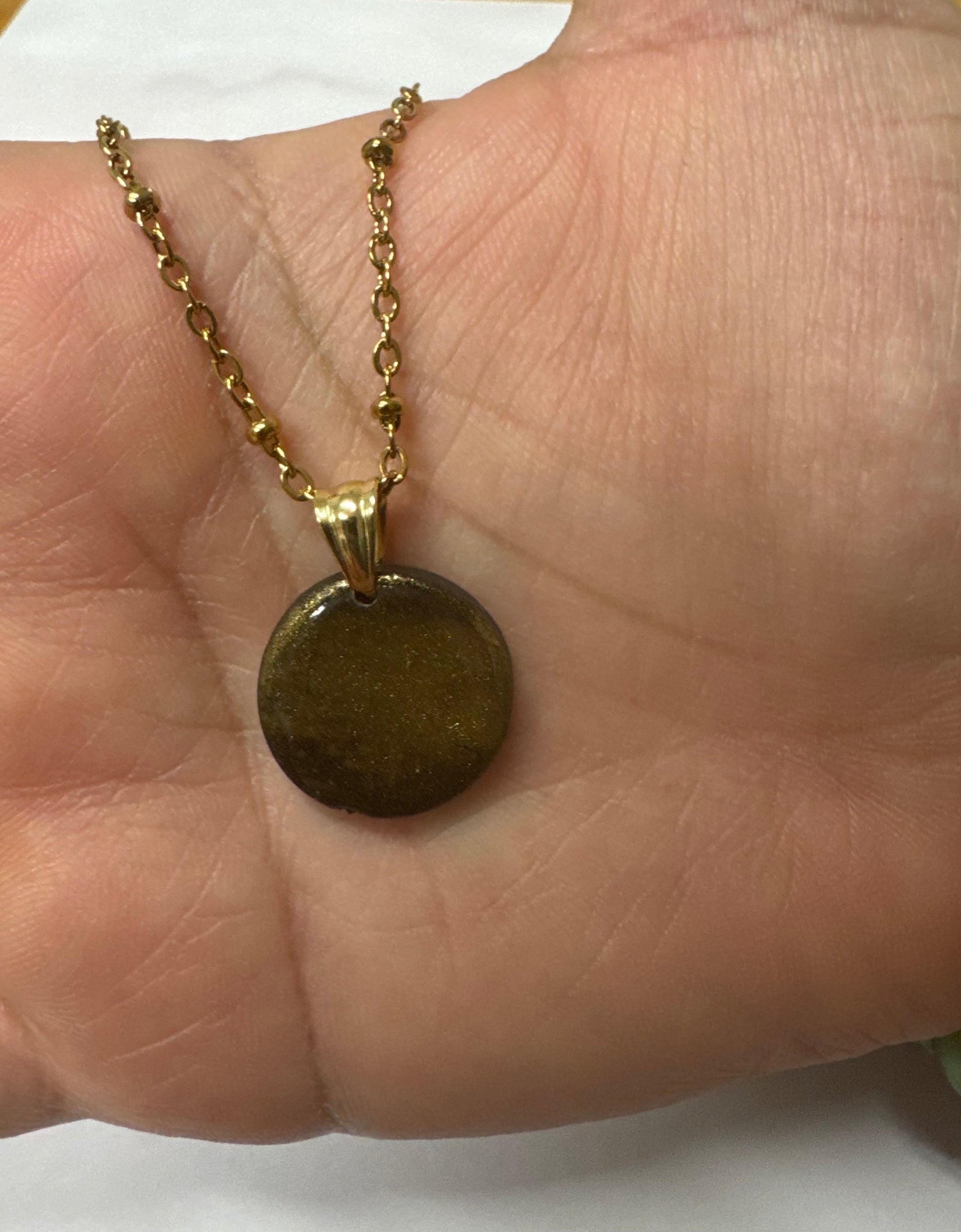 Ciondolo tondo nero con effetto brillantinato dorato appoggiato sul palmo di una mano.