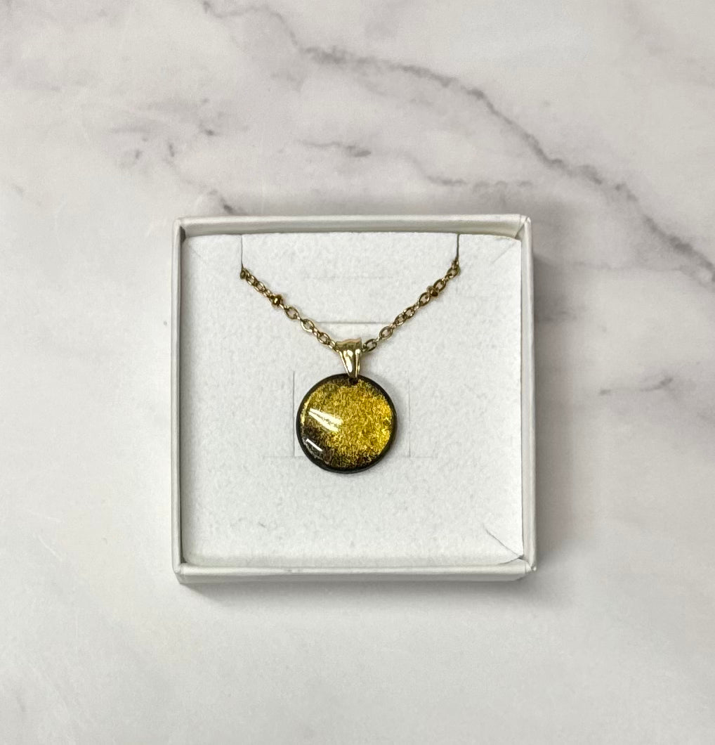 Ciondolo tondo nero con effetto brillantinato dorato, catena dorata, in scatolina regalo bianca aperta su sfondo marmo.