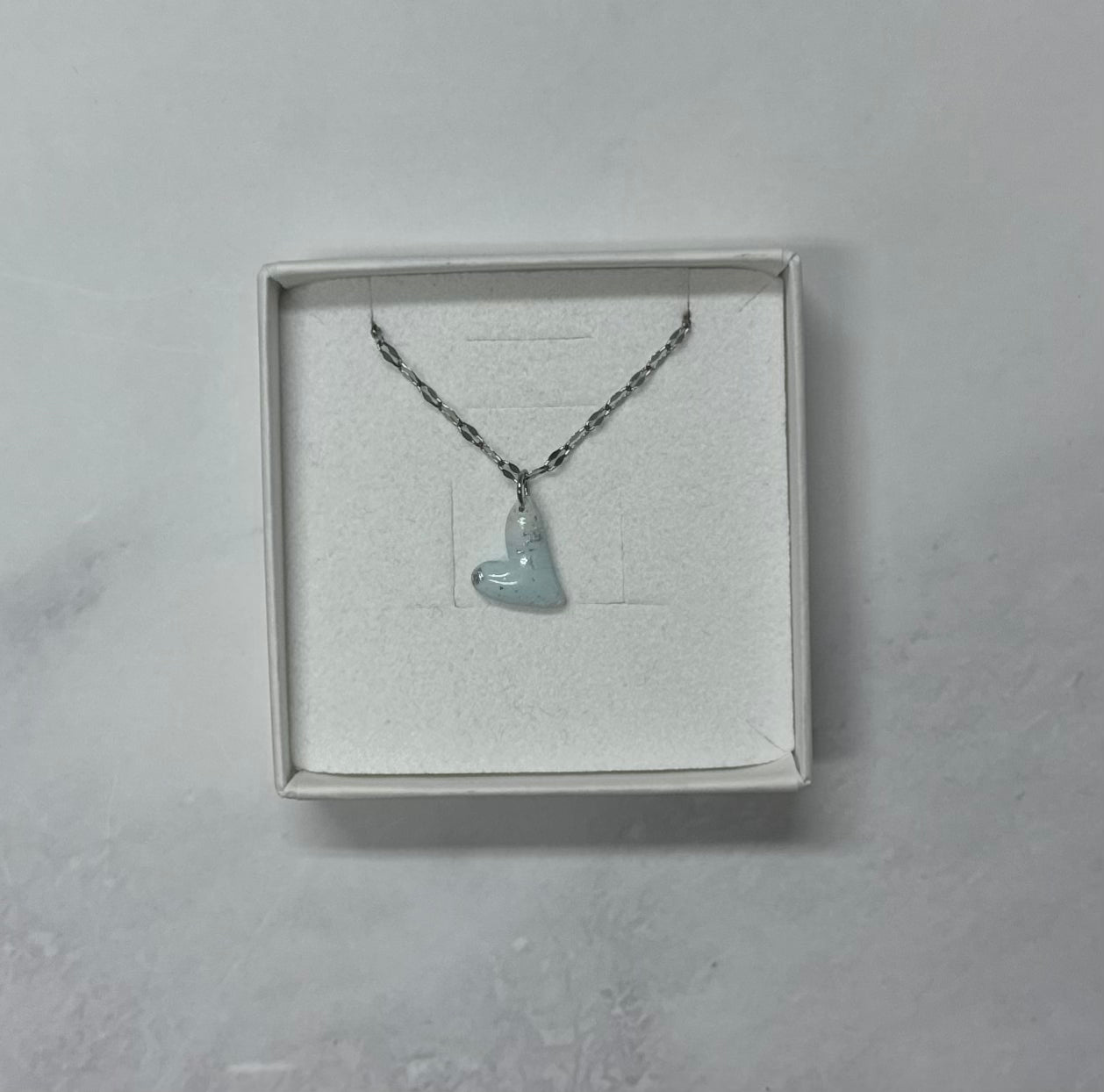 Ciondolo a cuore azzurro tenue con texture screziata, catena argentata, in scatolina regalo bianca aperta su sfondo marmo.