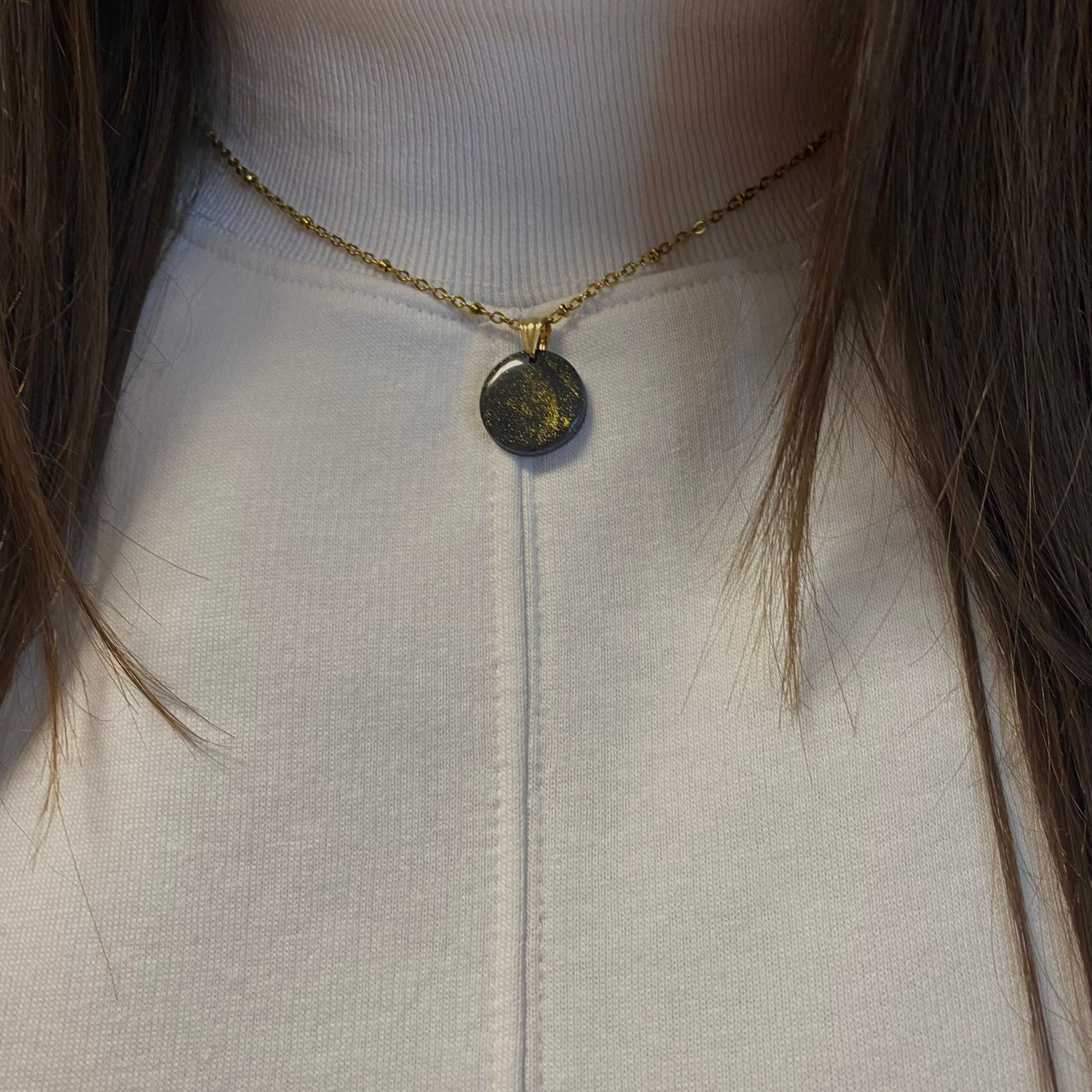 Primo piano della collana indossata su una maglietta bianca. Collana pendente con ciondolo rotondo in porcellana fredda, colore nero con dettagli in oro, con catenina dorata. 
Gioiello artigianale fatto a mano da persone cieche e ipovedenti.