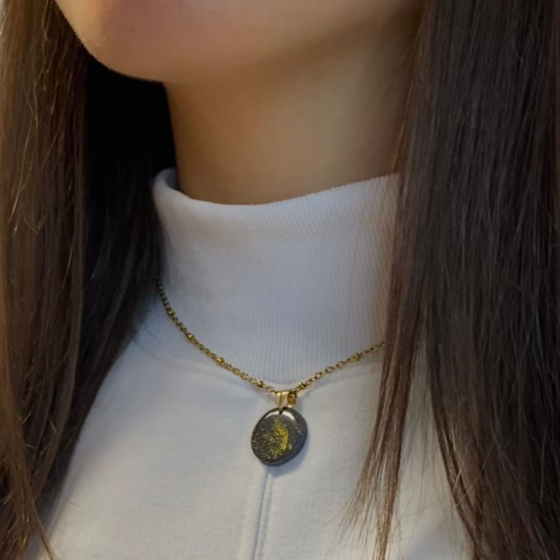 Primo piano laterale di una persona che indossa una collana pendente con ciondolo rotondo in porcellana fredda, colore nero con dettagli in oro, con catenina dorata. 
Gioiello artigianale fatto a mano da persone cieche e ipovedenti.

