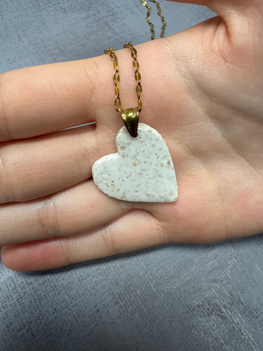 Mano che tiene nel palmo una collana con ciondolo a cuore bianco a pois in porcellana fredda bianca, sfumature dorate e catenina dorata. Gioiello artigianale fatto a mano da persone cieche e ipovedenti.