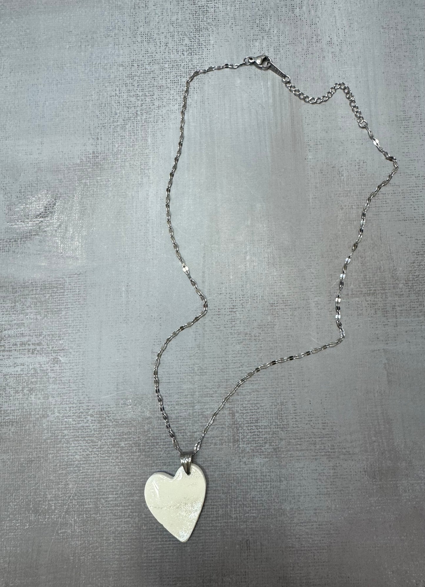 Collana con pendente cuore porcellana fredda, bianca con sfumature argentate e catenina argentata, vista a figura intera su sfondo grigio. Gioiello artigianale fatto a mano da persone cieche e ipovedenti.