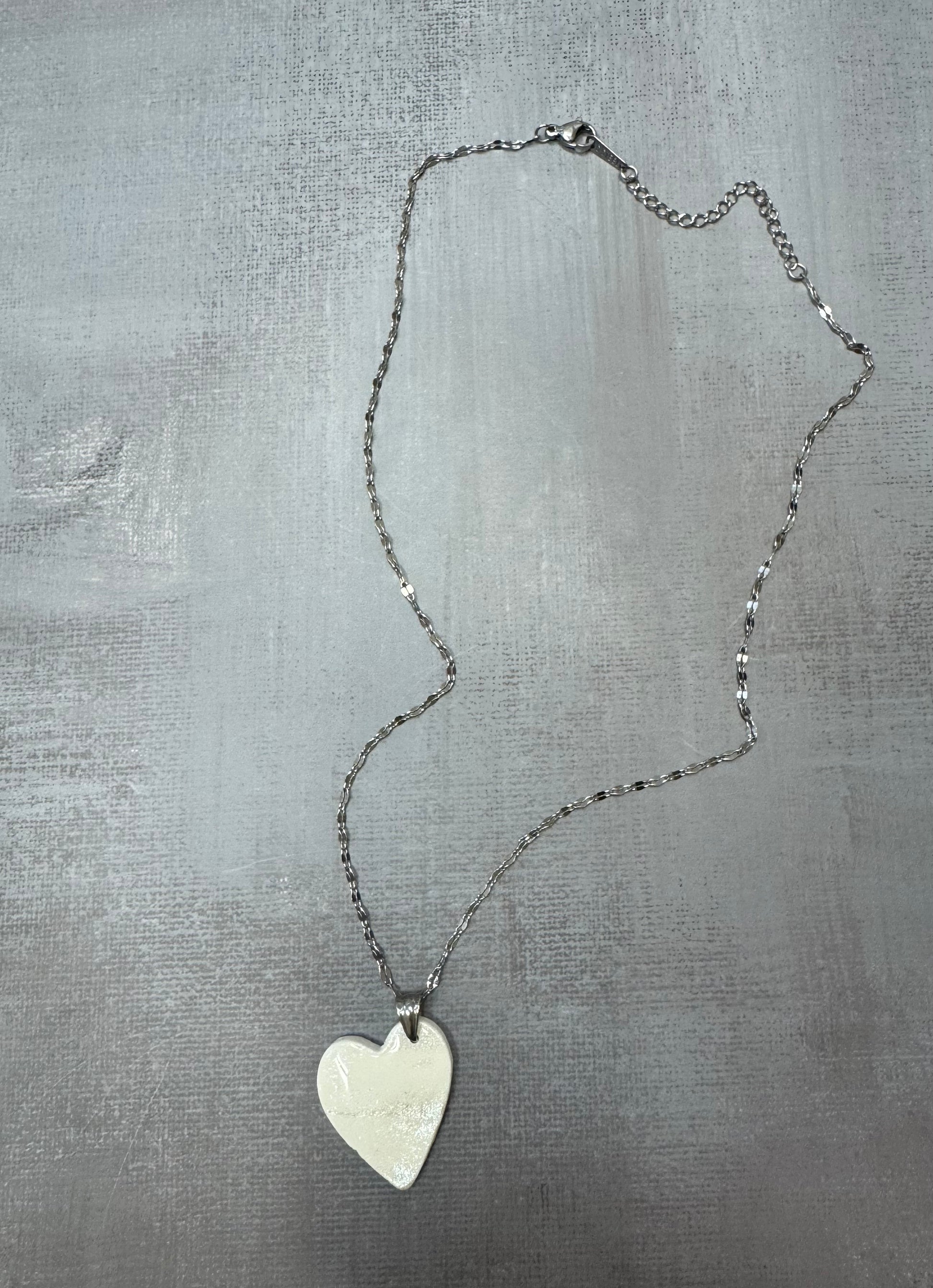 Collana con pendente cuore porcellana fredda, bianca con sfumature argentate e catenina argentata, vista a figura intera su sfondo grigio. Gioiello artigianale fatto a mano da persone cieche e ipovedenti.