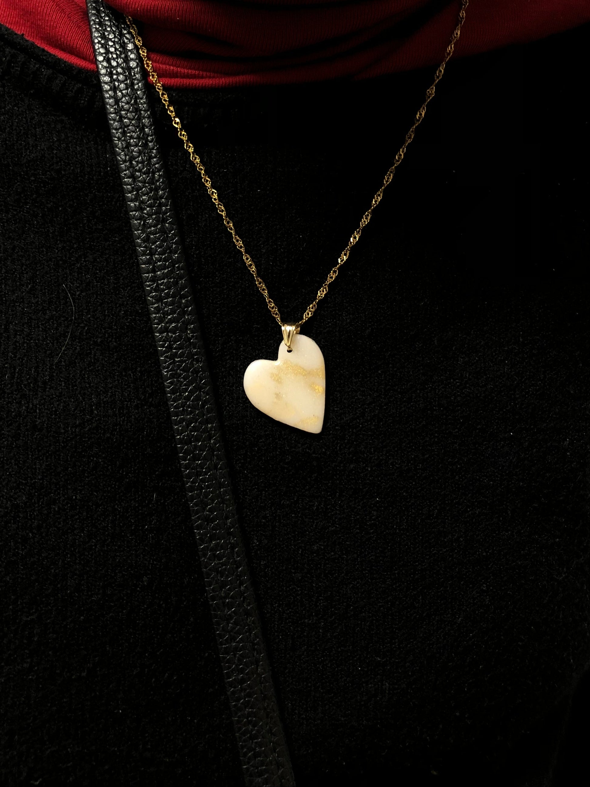 Primo piano della collana indossata su una maglietta nera. Collana pendente con ciondolo a forma di cuore in porcellana fredda, colore bianco e oro, con catenina dorata. 
Gioiello artigianale fatto a mano da persone cieche e ipovedenti.