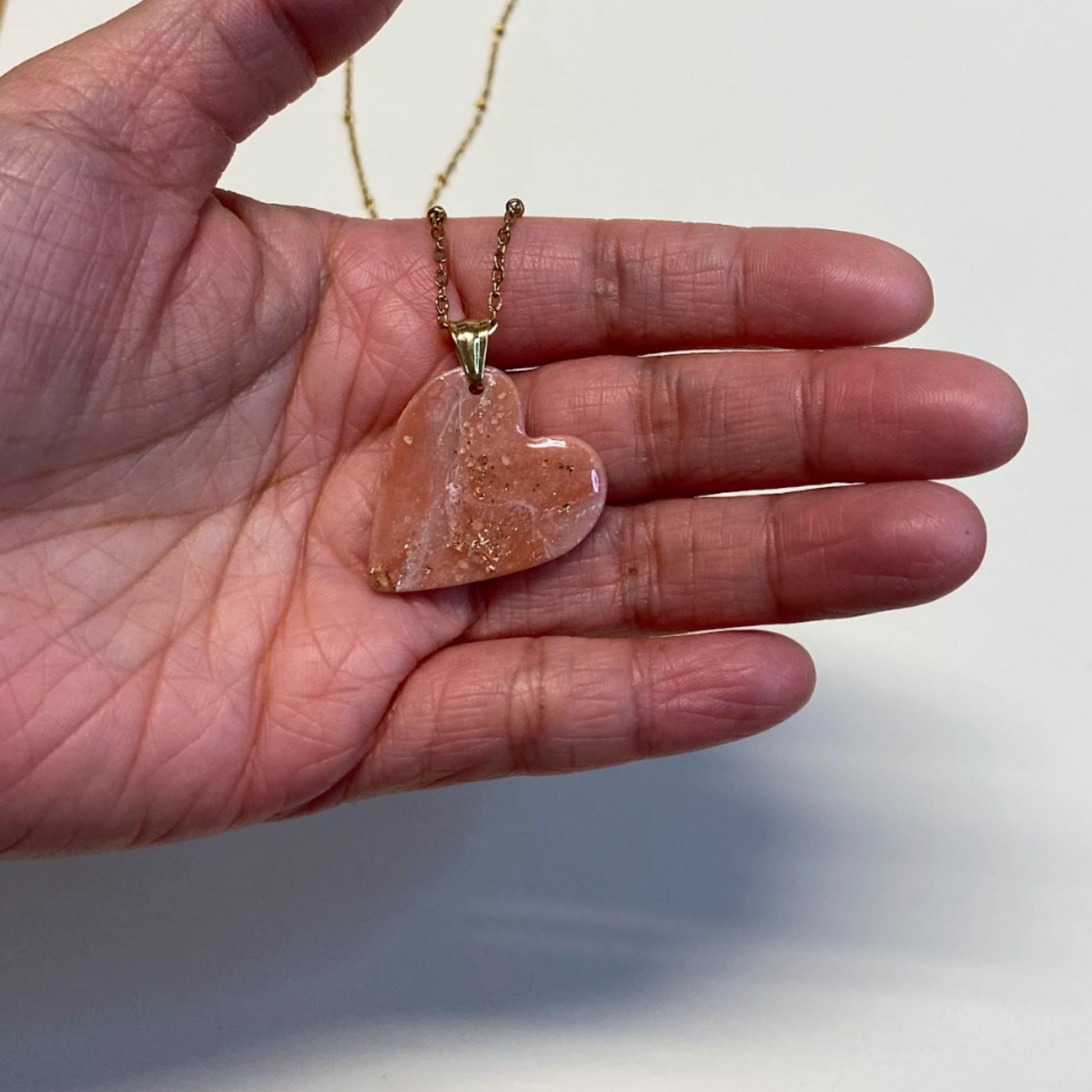 Collana pendente con ciondolo a forma di cuore, in porcellana fredda, colore rosa e bianco, effetto marmo, con catenina dorata. 
Gioiello artigianale fatto a mano da persone cieche e ipovedenti. 
La collana viene tenuta in mano. 