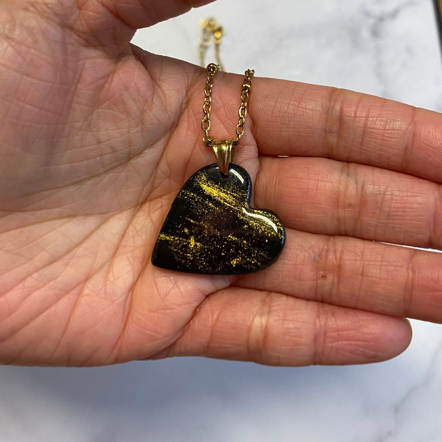 Collana pendente con ciondolo a forma di cuore in porcellana fredda, colore nero con dettagli in oro, effetto marmo, con catenina dorata. 
Gioiello artigianale fatto a mano da persone cieche e ipovedenti. 
La collana viene tenuta in mano. 