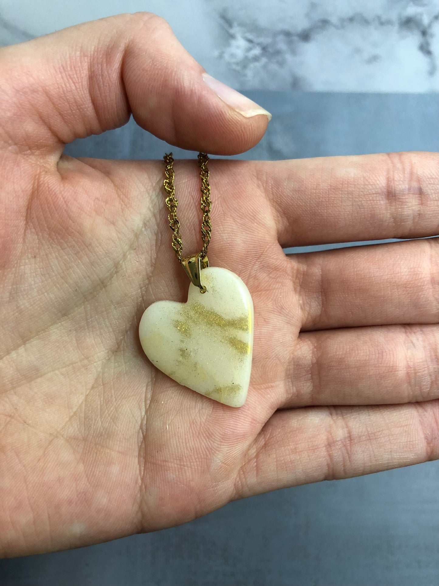 Collana pendente con ciondolo a forma di cuore in porcellana fredda, colore bianco con dettagli in oro, con catenina dorata. 
Gioiello artigianale fatto a mano da artigiani ciechi e ipovedenti. 
La collana viene tenuta in mano. 
