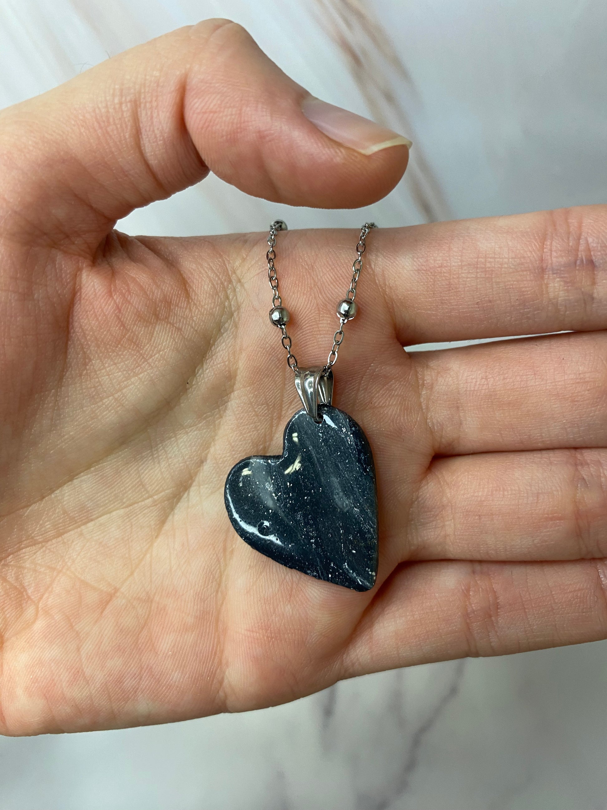 Collana pendente con ciondolo a forma di cuore in porcellana fredda, colore nero con dettagli in argento, con catenina argentata. 
Gioiello artigianale fatto a mano da artigiani ciechi e ipovedenti. 
La collana viene tenuta in mano. 