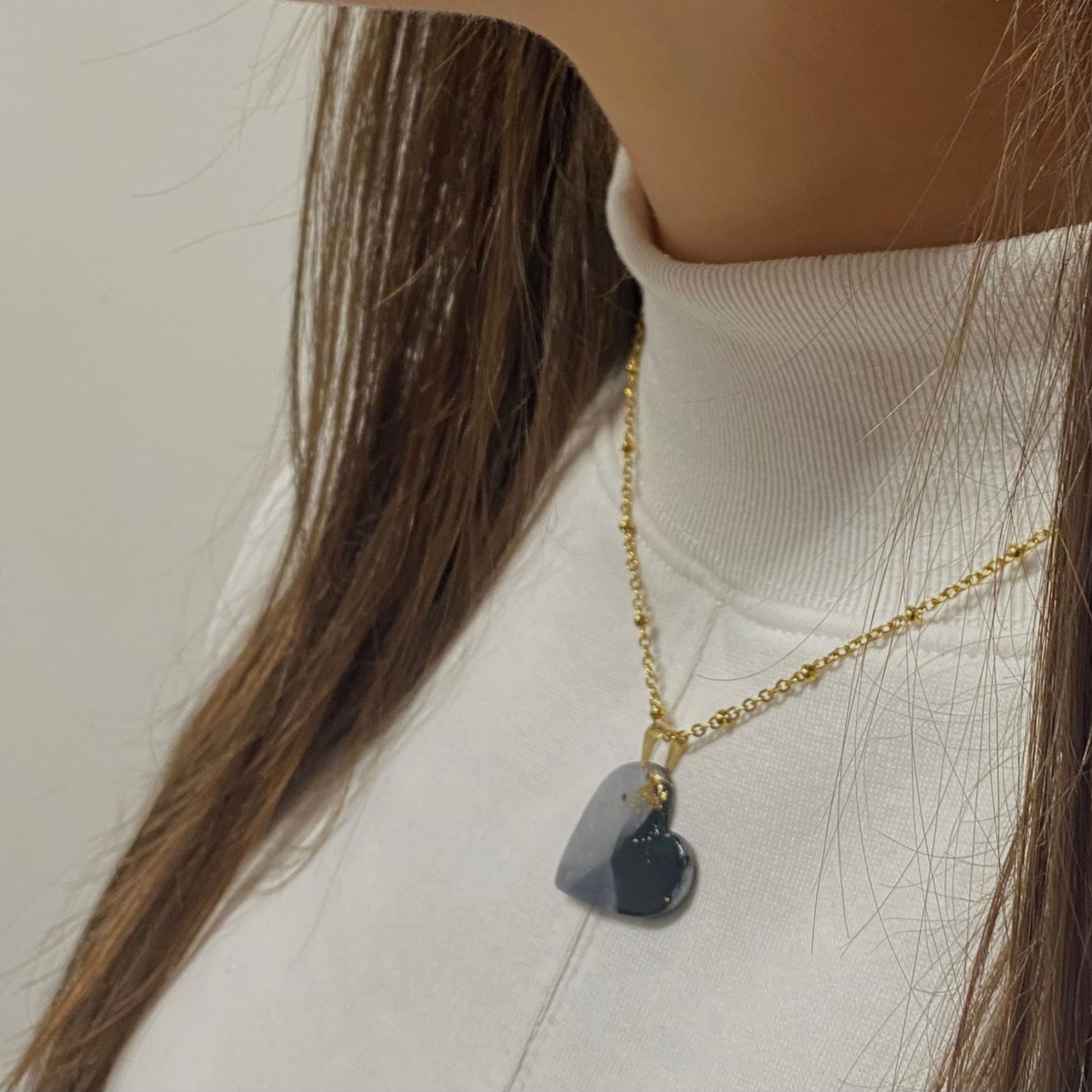 Primo piano laterale di una persona che indossa una collana pendente con ciondolo a forma di cuore, in porcellana fredda, colore nero e bianco, effetto marmo sfumato, con catenina dorata. Gioiello artigianale fatto a mano da persone cieche e ipovedenti. 