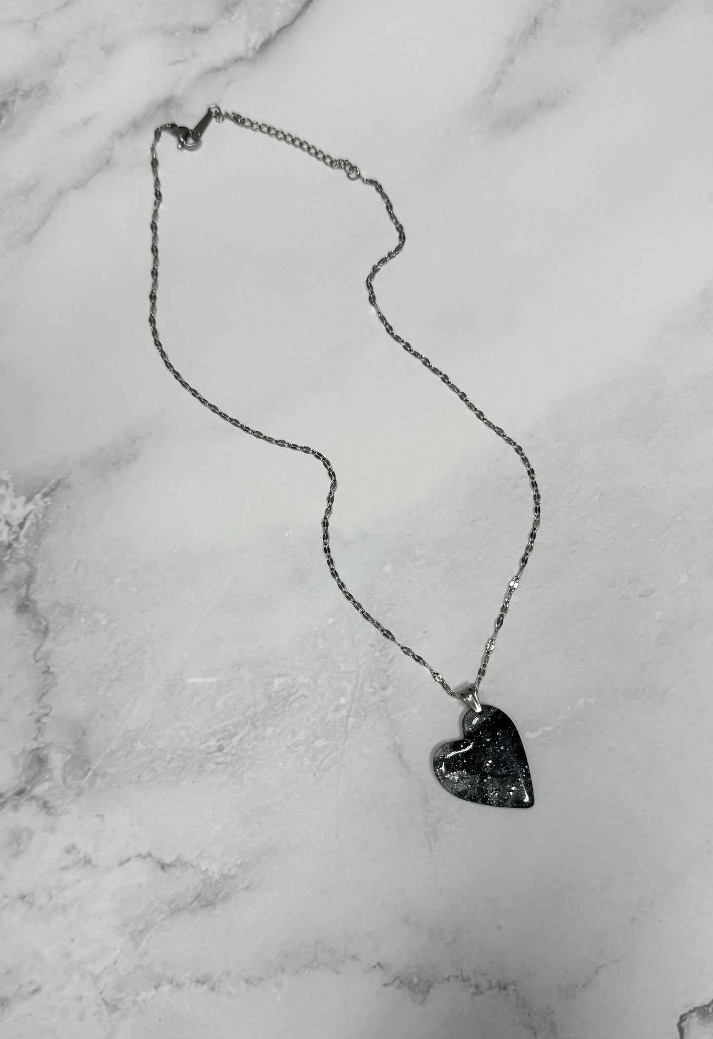 Vista completa della collana pendente su sfondo marmo, con cuore nero e argento effetto marmo in porcellana fredda e catenina argentata. 