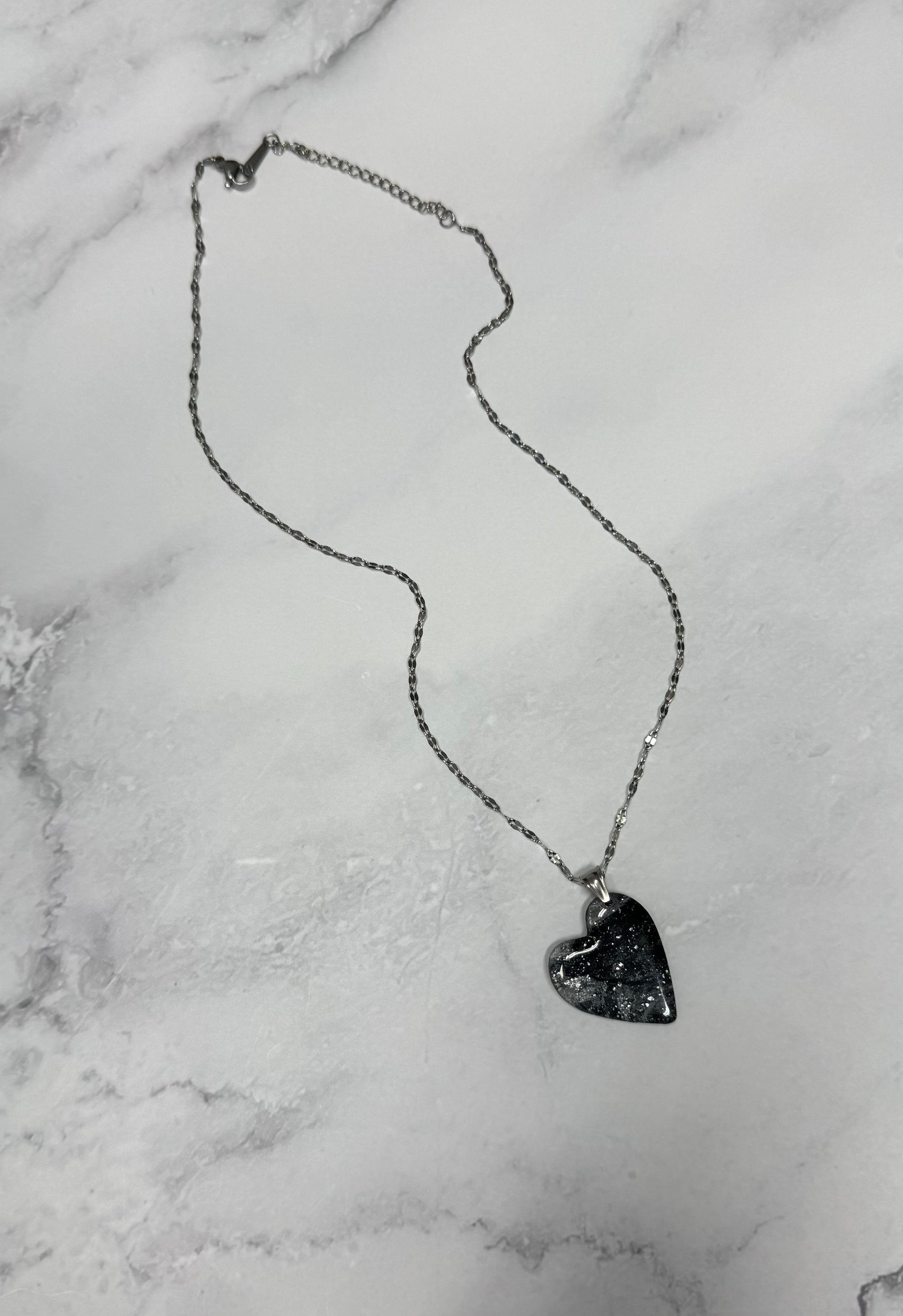 Vista completa della collana pendente su sfondo marmo, con cuore nero e argento effetto marmo in porcellana fredda e catenina argentata. 