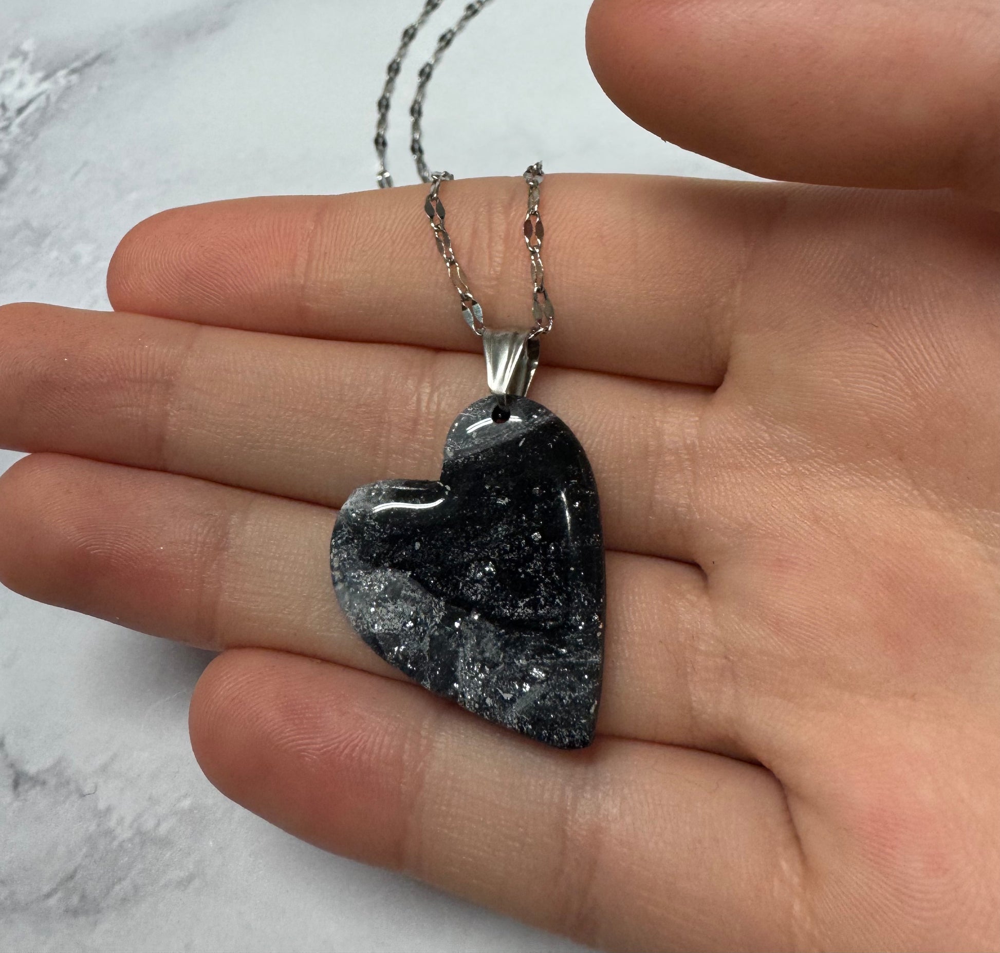 Collana pendente con ciondolo a forma di cuore in porcellana fredda, colore nero con sfumature argentate, effetto marmo, con catenina argentata. Gioiello artigianale fatto a mano da persone cieche e ipovedenti. La collana viene tenuta in mano.