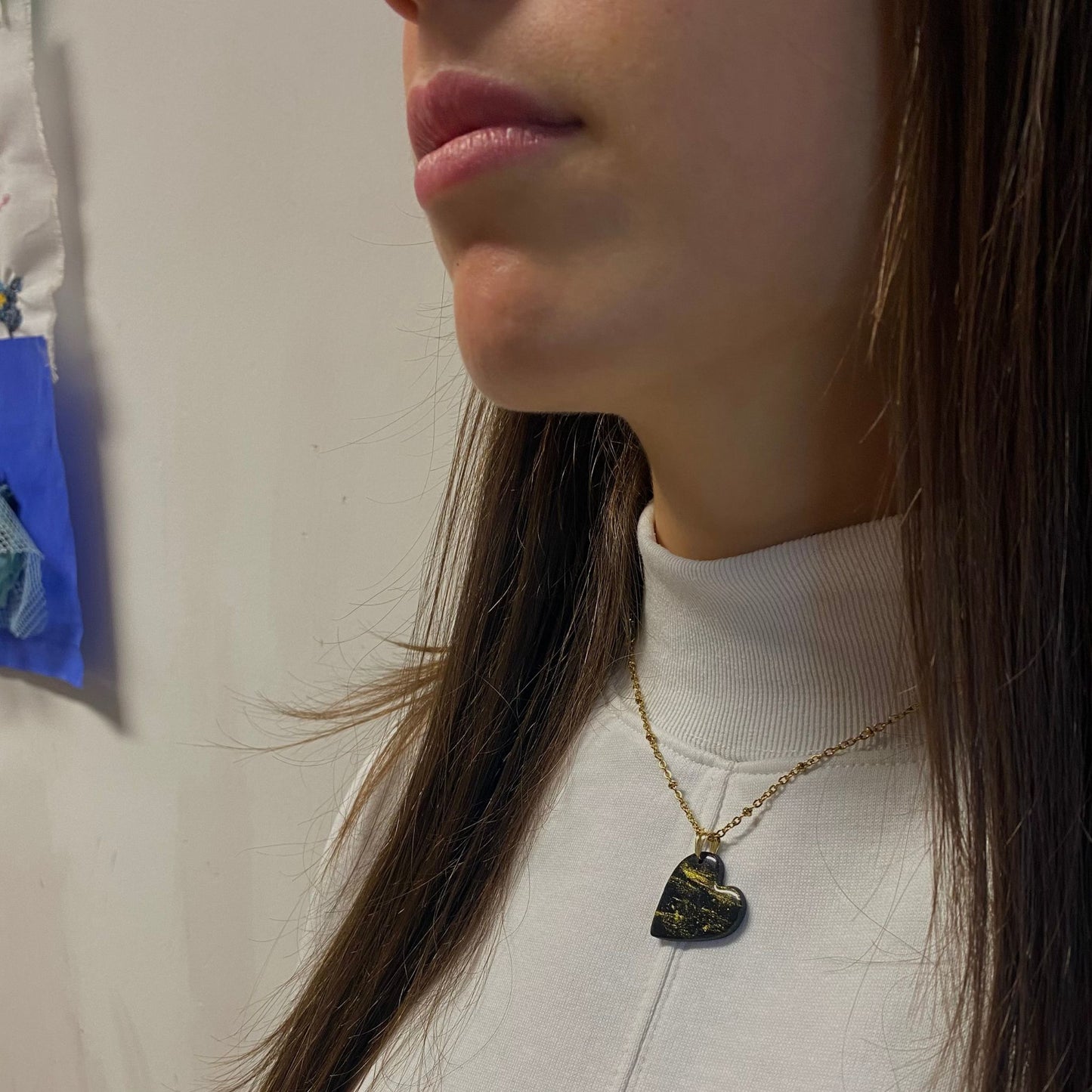 Primo piano laterale di una persona che indossa una collana pendente con ciondolo a forma di cuore, in porcellana fredda, colore nero con dettagli in oro, effetto marmo, con catenina dorata. Gioiello artigianale fatto a mano da persone cieche e ipovedenti. 