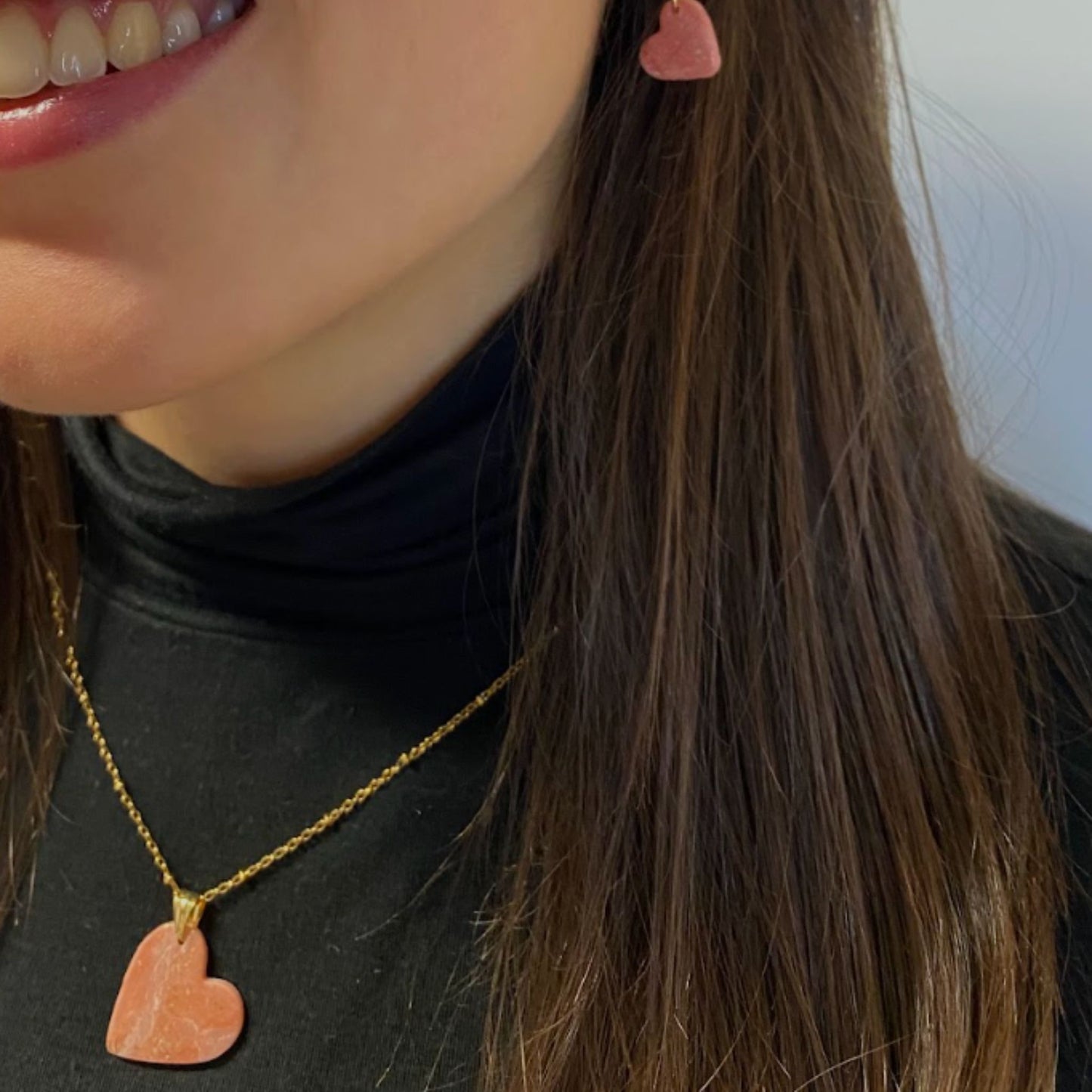Primo piano di una persona sorridente, ripresa di lato, che indossa la collana pendente con ciondolo a forma di cuore, in porcellana fredda, colore rosa e bianco, con dettagli oro, effetto marmo, con catenina dorata. 
Gioiello artigianale fatto a mano da persone cieche e ipovedenti.