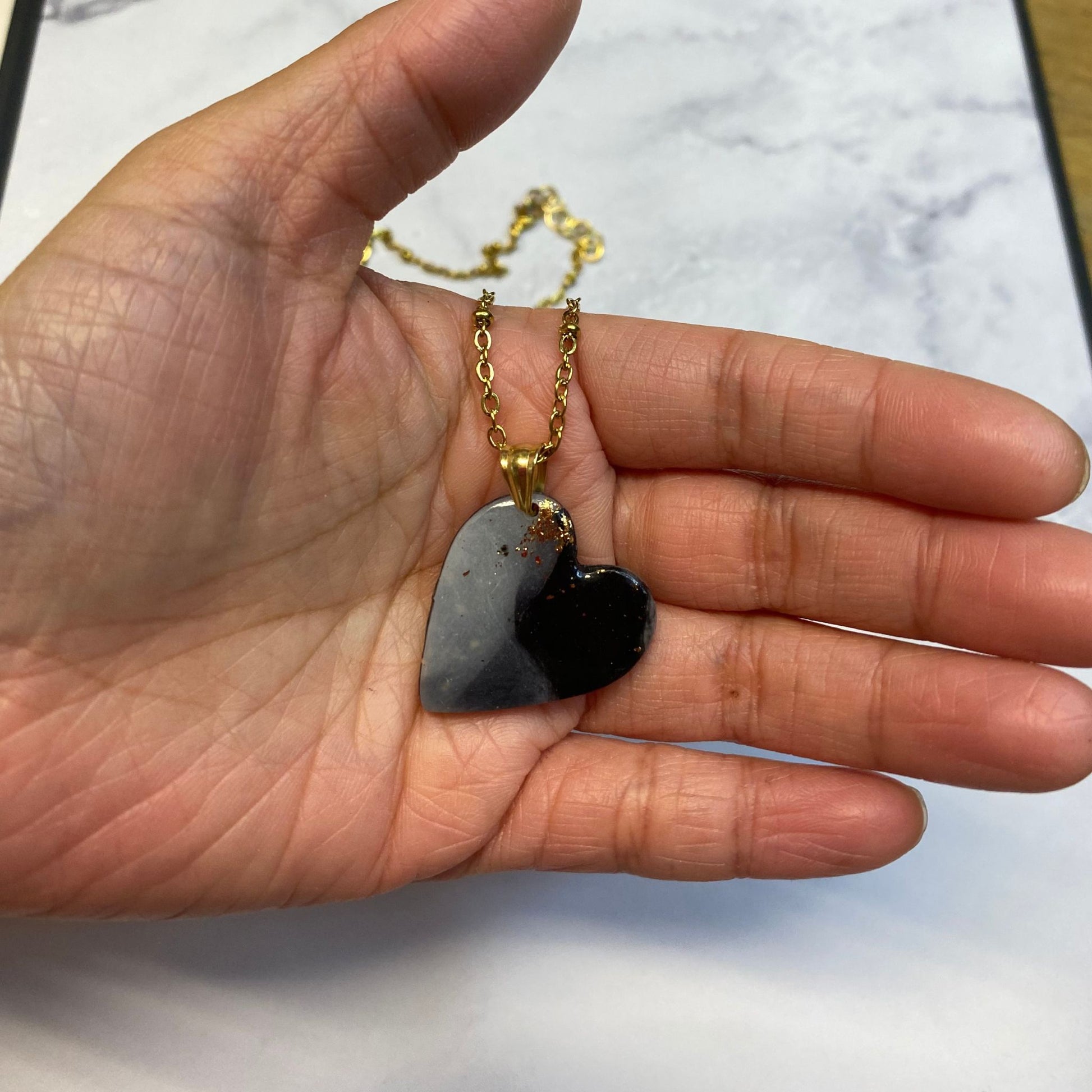 Collana pendente con ciondolo a forma di cuore in porcellana fredda, colore nero e bianco, effetto marmo sfumato, con catenina dorata. 
Gioiello artigianale fatto a mano da persone cieche e ipovedenti. 
La collana viene tenuta in mano. 