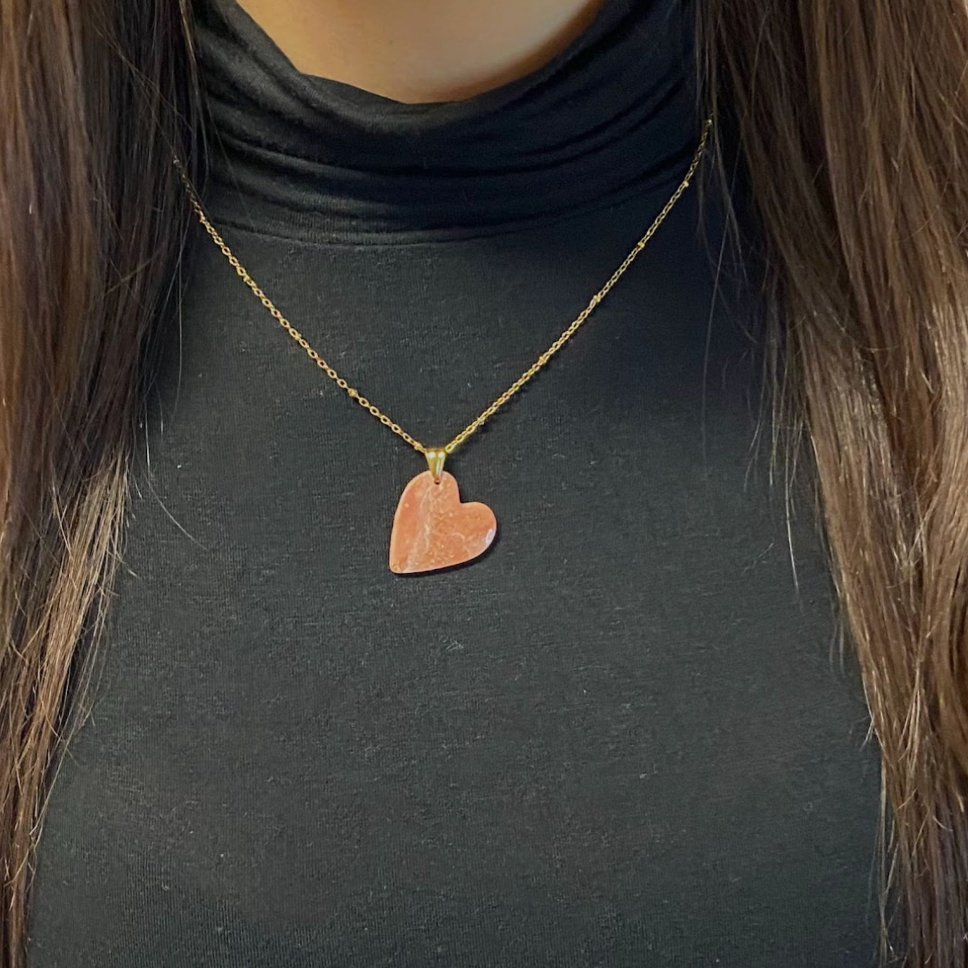 Primo piano della collana indossata su un dolcevita nero. Collana pendente con ciondolo a forma di cuore, in porcellana fredda, colore rosa e bianco con dettagli oro, effetto marmo, con catenina dorata. Gioiello artigianale fatto a mano da persone cieche e ipovedenti. 