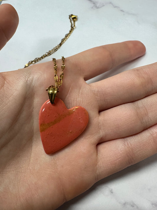 Collana con cuore rosso in porcellana fredda e dettagli dorati, sul palmo di una mano. Gioiello realizzato da artigiani ciechi e ipovedenti.