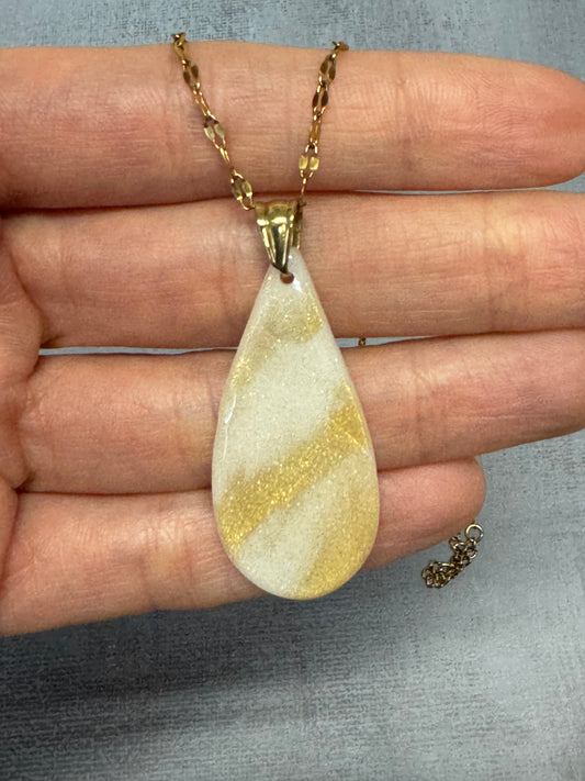 Collana pendente con ciondolo a goccia in porcellana fredda, bianco con sfumature dorate. Gioiello artigianale fatto a mano da persone cieche e ipovedenti. La collana viene tenuta in mano.