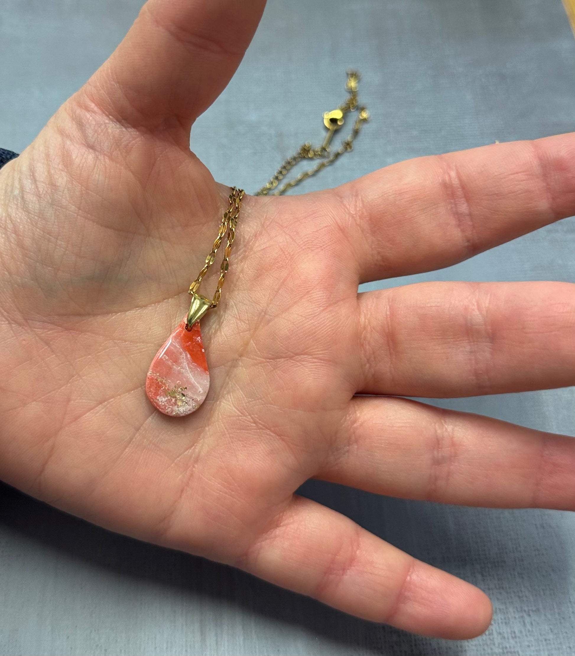 Collana con ciondolo a goccia, rosa marmorizzato in porcellana fredda con sfumature e dettagli dorati, tenuta sul palmo di una mano. Gioiello realizzato da artigiani ciechi e ipovedenti.