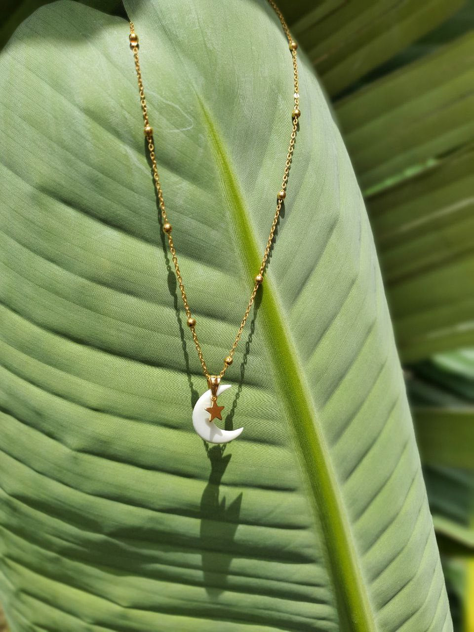 Collana Myra Lab La Luna con pendente in porcellana fredda bianca e stellina dorata, gioiello artigianale inclusivo creato da persone cieche e ipovedenti, fotografato su foglia verde