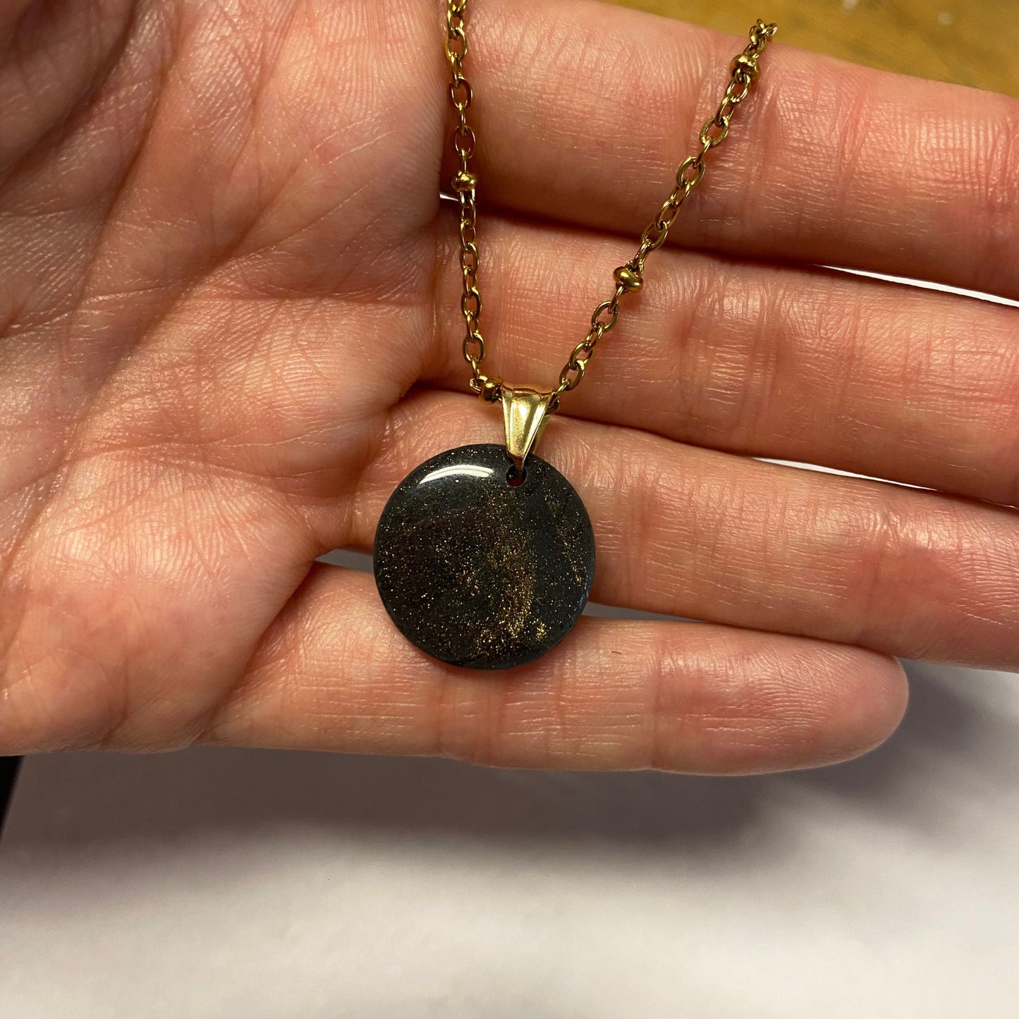 Collana pendente con ciondolo rotondo in porcellana fredda, colore nero con dettagli in oro, con catenina dorata. 
Gioiello artigianale fatto a mano da artigiani ciechi e ipovedenti. 
La collana viene tenuta in mano. 