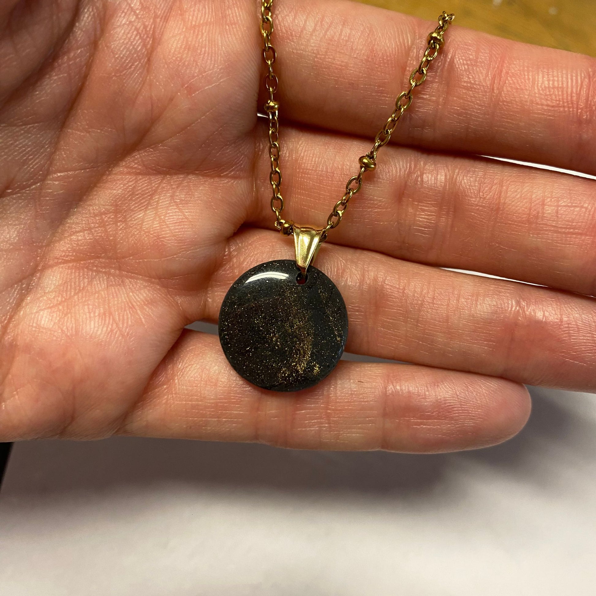 Collana pendente con ciondolo rotondo in porcellana fredda, colore nero con dettagli in oro, con catenina dorata. 
Gioiello artigianale fatto a mano da artigiani ciechi e ipovedenti. 
La collana viene tenuta in mano. 