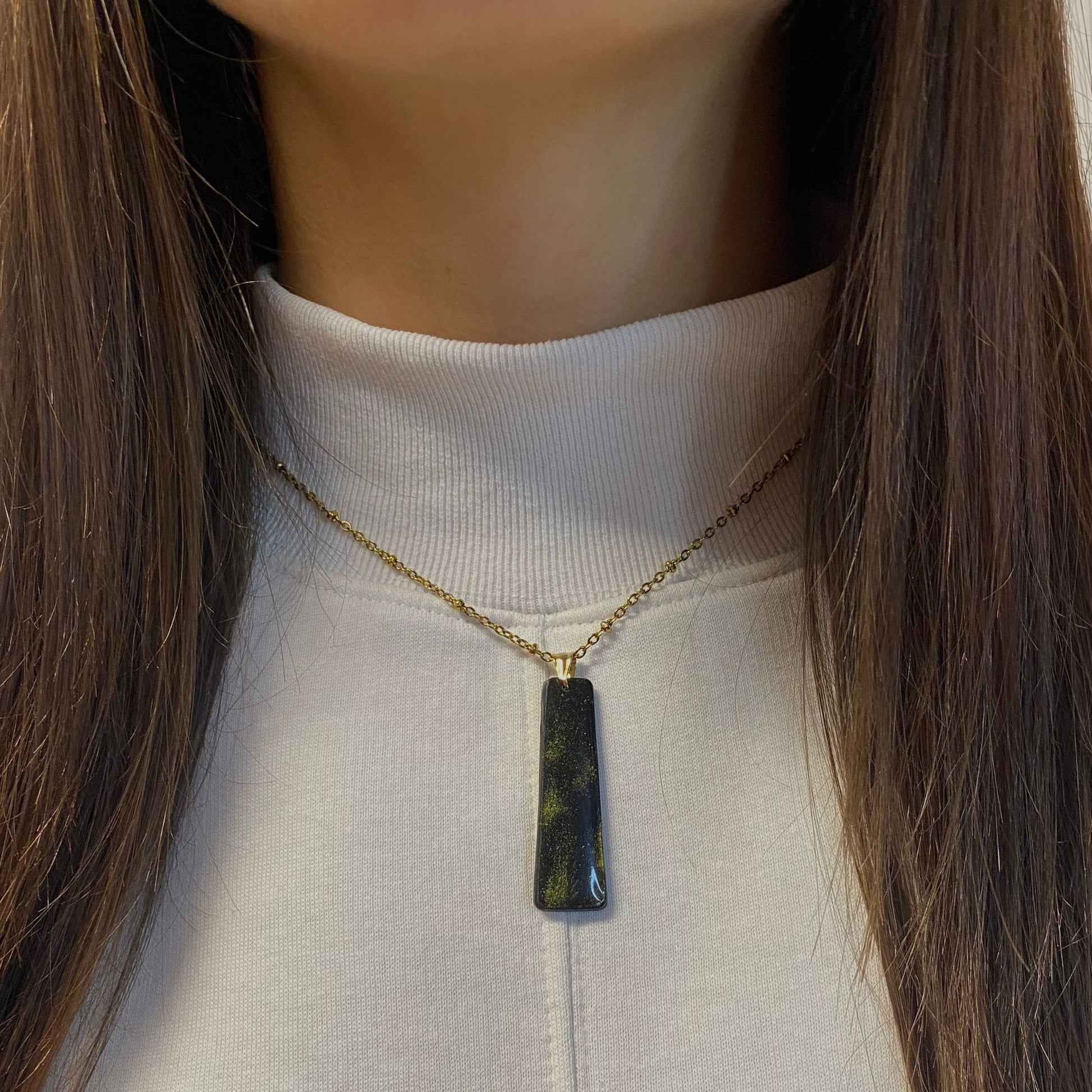Primo piano della collana indossata su una maglietta bianca. Collana pendente con ciondolo rettangolare allungato in porcellana fredda, colore nero e oro, con catenina dorata. 
Gioiello artigianale fatto a mano da persone cieche e ipovedenti.