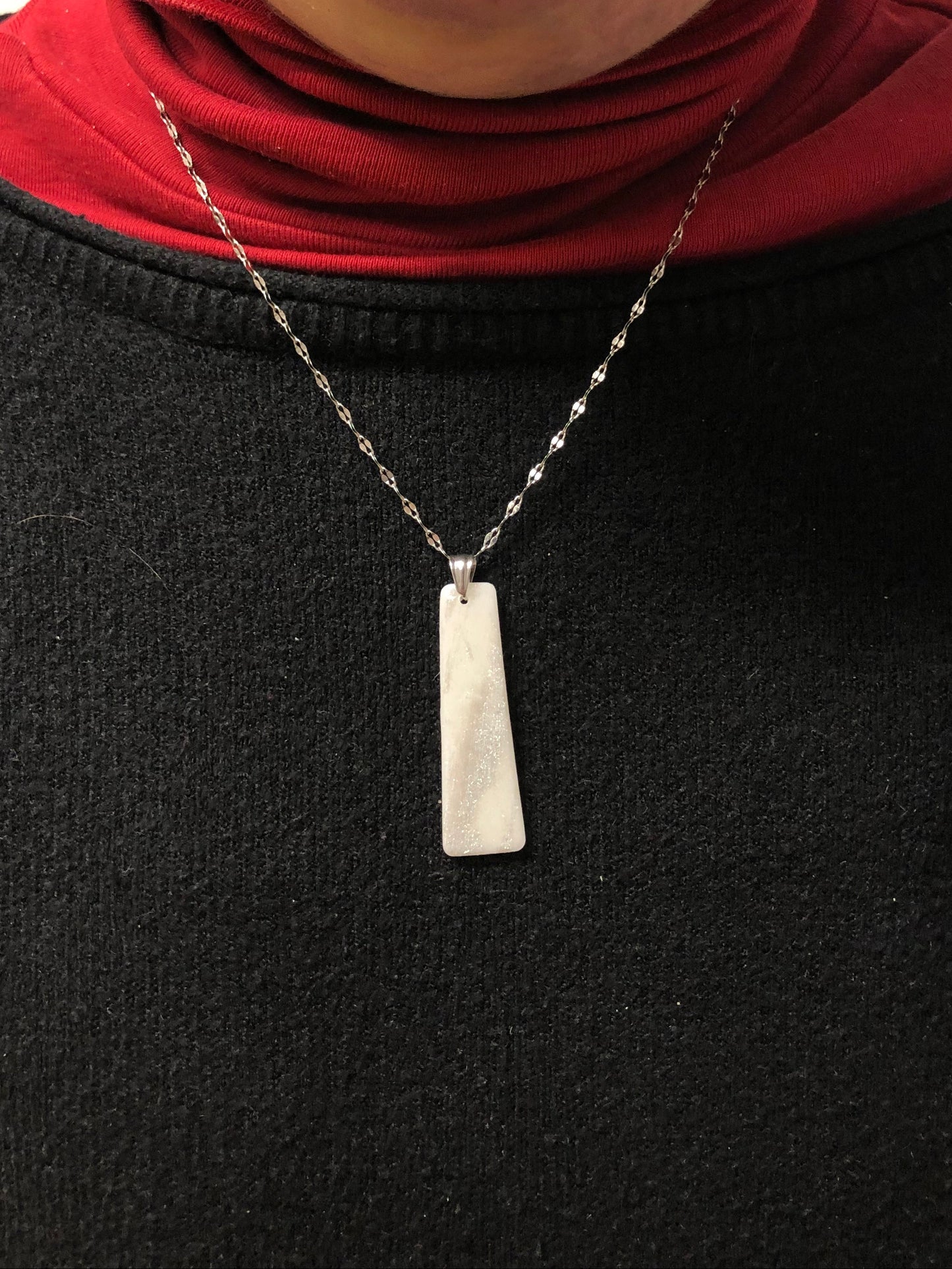 Primo piano della collana indossata su una maglietta nera. Collana pendente con ciondolo rettangolare in porcellana fredda, colore bianco e argento, con catenina argentata. 
Gioiello artigianale fatto a mano da persone cieche e ipovedenti.