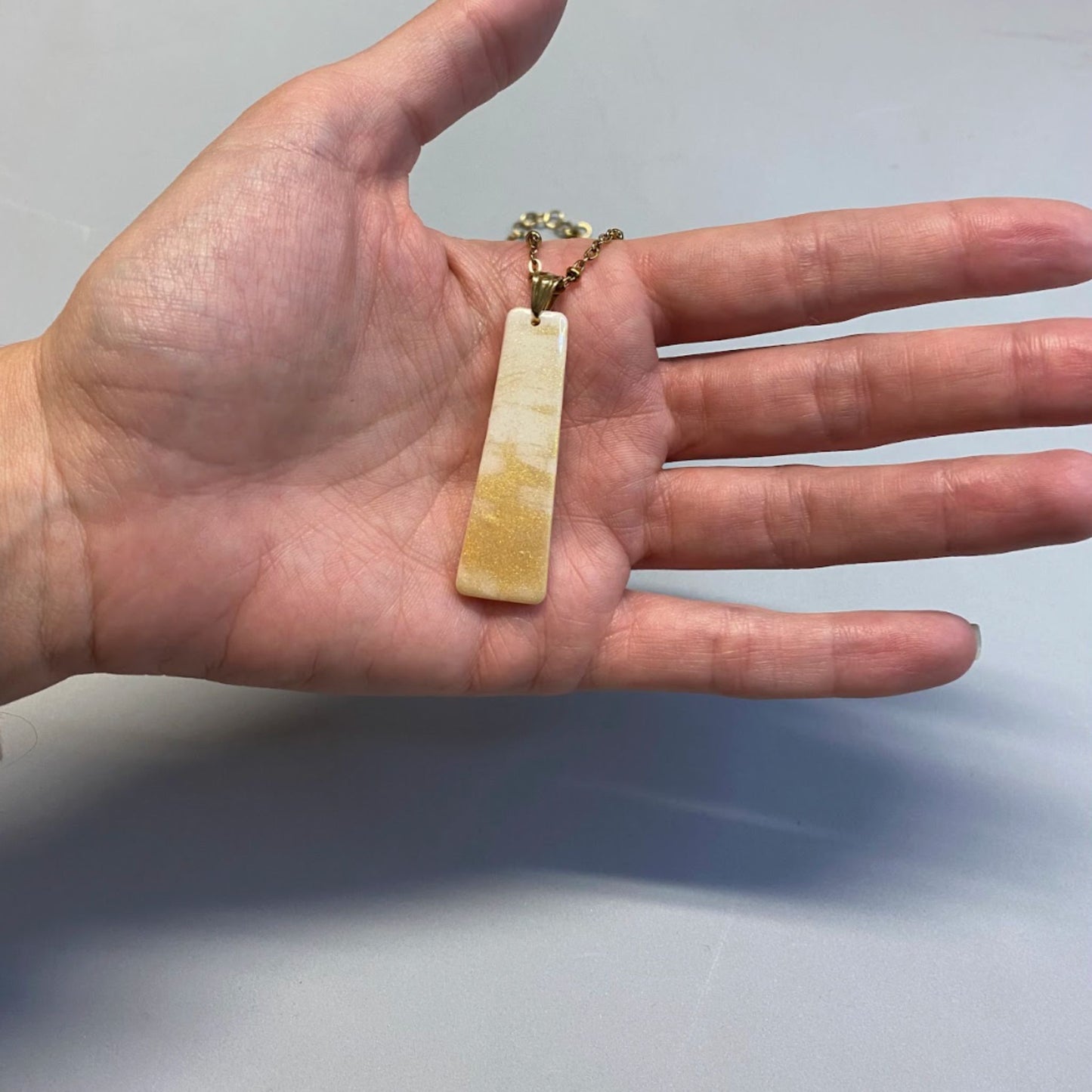 Collana pendente con ciondolo rettangolare allungato in porcellana fredda, colore bianco e oro, effetto marmo, con catenina dorata. 
Gioiello artigianale fatto a mano da persone cieche e ipovedenti. 
La collana viene tenuta in mano. 