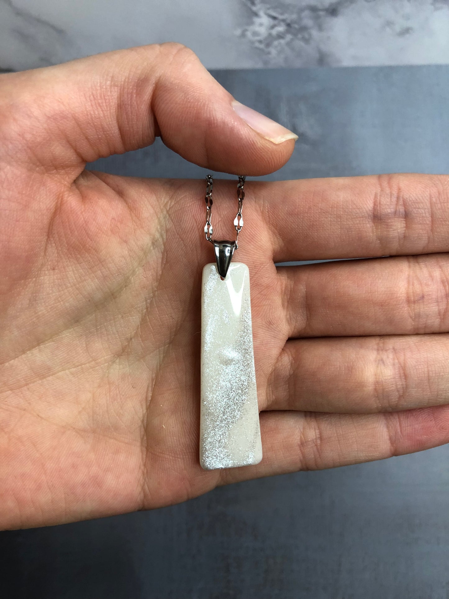 Collana pendente con ciondolo rettangolare in porcellana fredda, colore bianco con dettagli in argento, con catenina argentata. 
Gioiello artigianale fatto a mano da artigiani ciechi e ipovedenti. 
La collana viene tenuta in mano. 