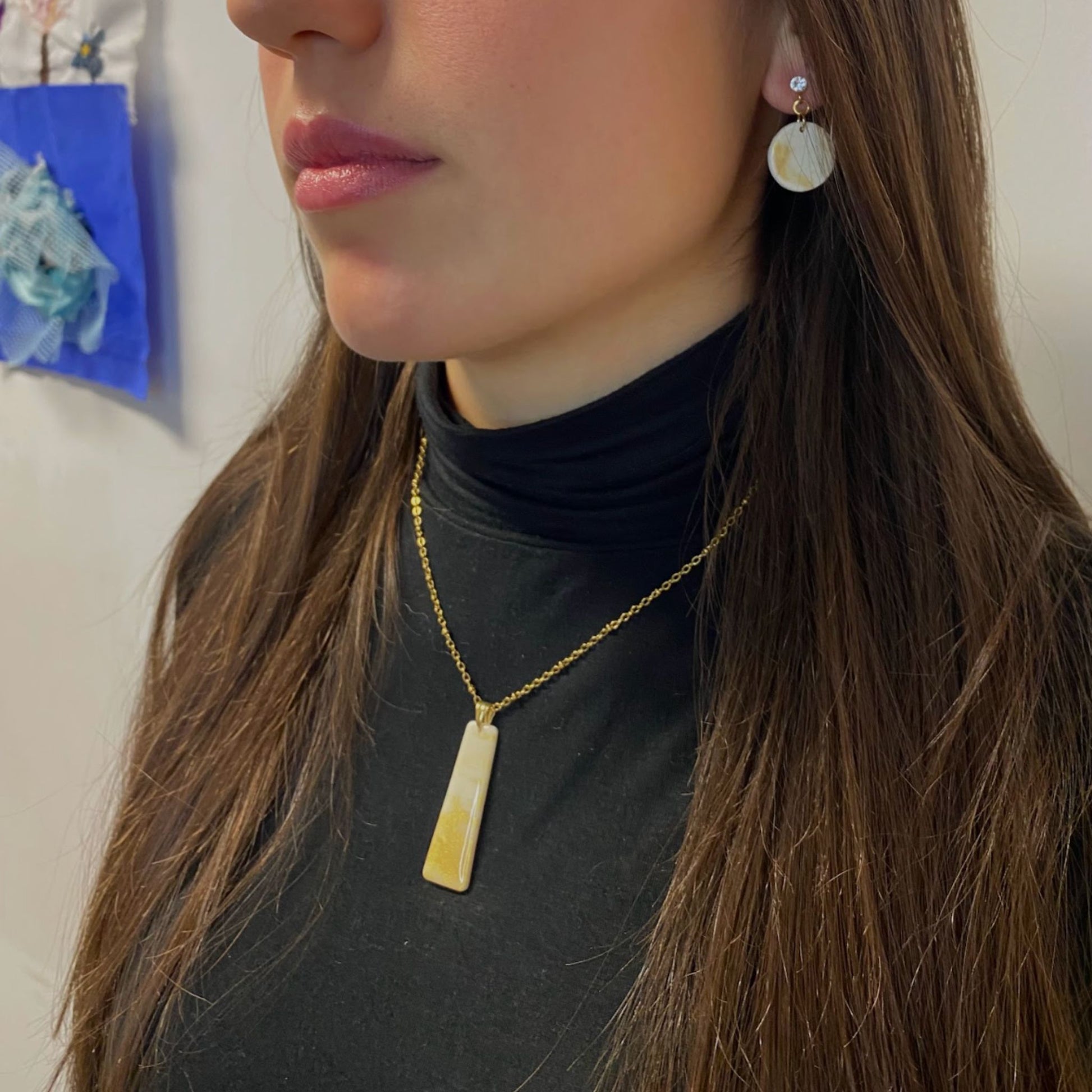 Persona ritratta di profilo con parte del viso visibile. Indossa una collana pendente con ciondolo rettangolare allungato in porcellana fredda, colore bianco e oro, effetto marmo, con catenina dorata. Gioielli realizzati a mano da artigiani ciechi e ipovedenti. 
