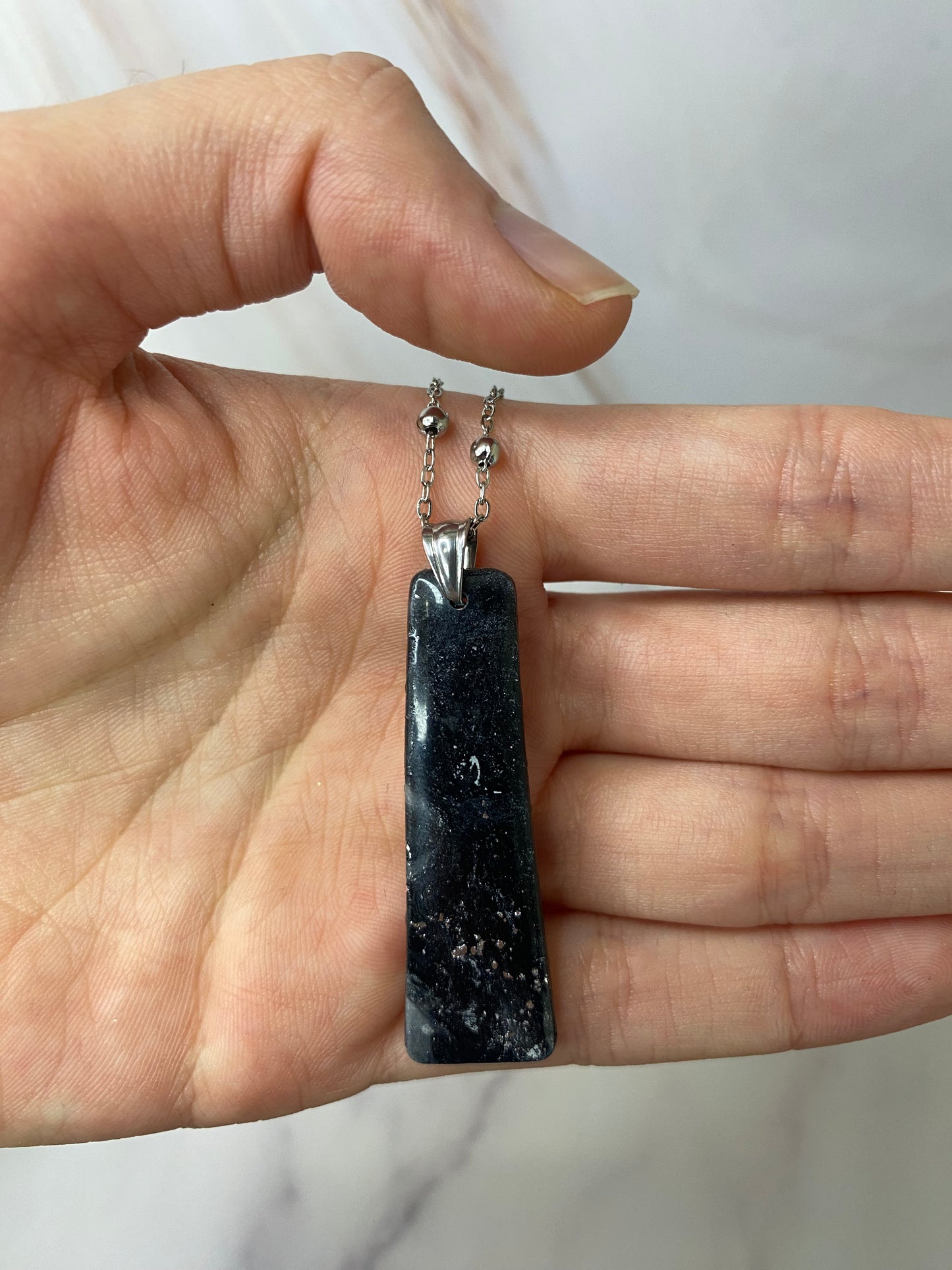 Collana pendente con ciondolo rettangolare in porcellana fredda, colore nero con dettagli in argento, con catenina argentata. 
Gioiello artigianale fatto a mano da artigiani ciechi e ipovedenti. 
La collana viene tenuta in mano. 